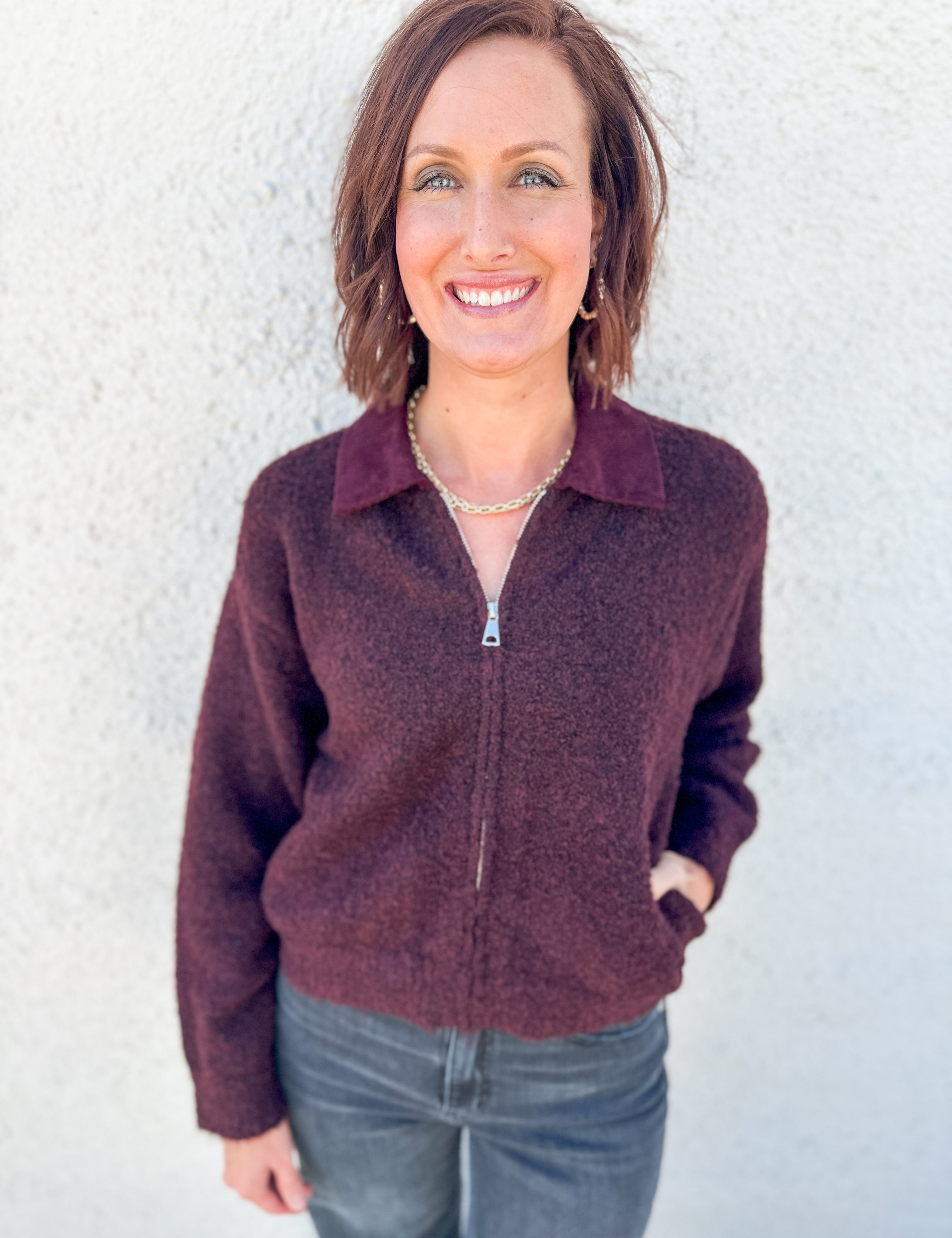 A smiling woman with short brown hair wearing a heathered dark plum-brown zip-up quarter-collar sweater with a corduroy collar by Kut from the Kloth and gray jeans, standing against a light textured wall.