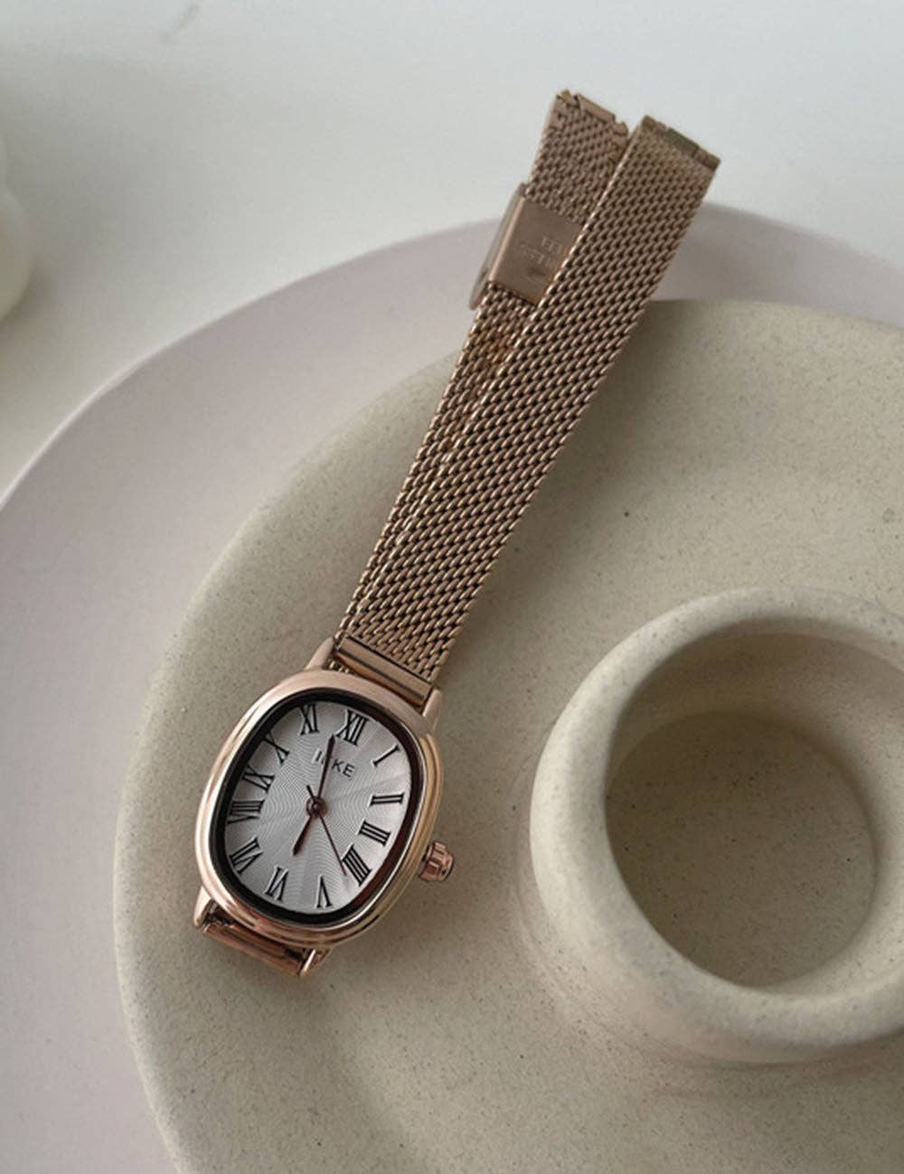 A vintage-style wristwatch with an oval  gold case, a white dial with black Roman numerals, and a matching gold mesh wrist strap, resting on a textured ceramic dish.