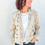 A woman wearing a beige and gray jacquard print jacket by Free People over a white t-shirt and dark gray jeans, standing against a light gray textured wall.