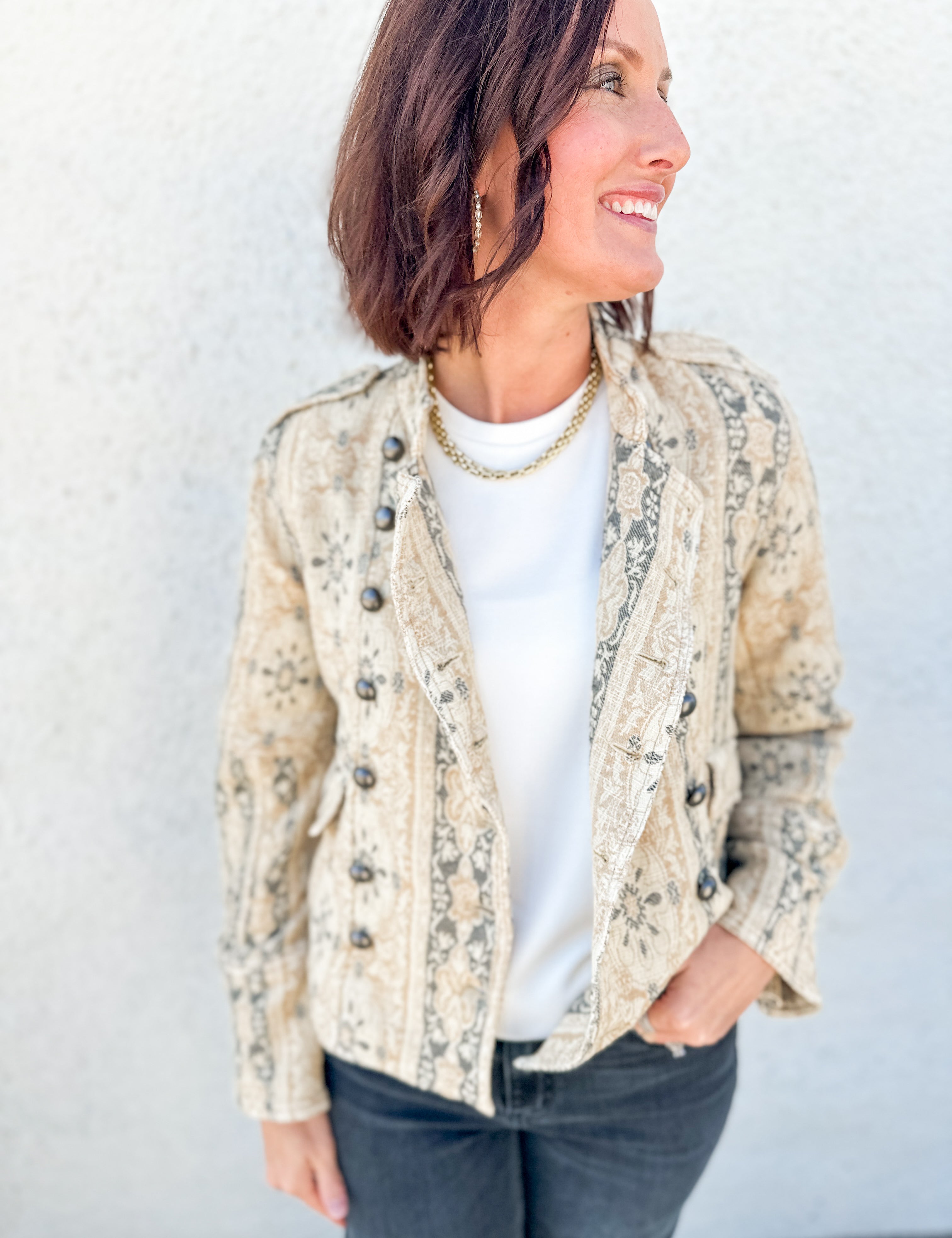 A woman wearing a beige and gray jacquard print jacket by Free People over a white t-shirt and dark gray jeans, standing against a light gray textured wall.