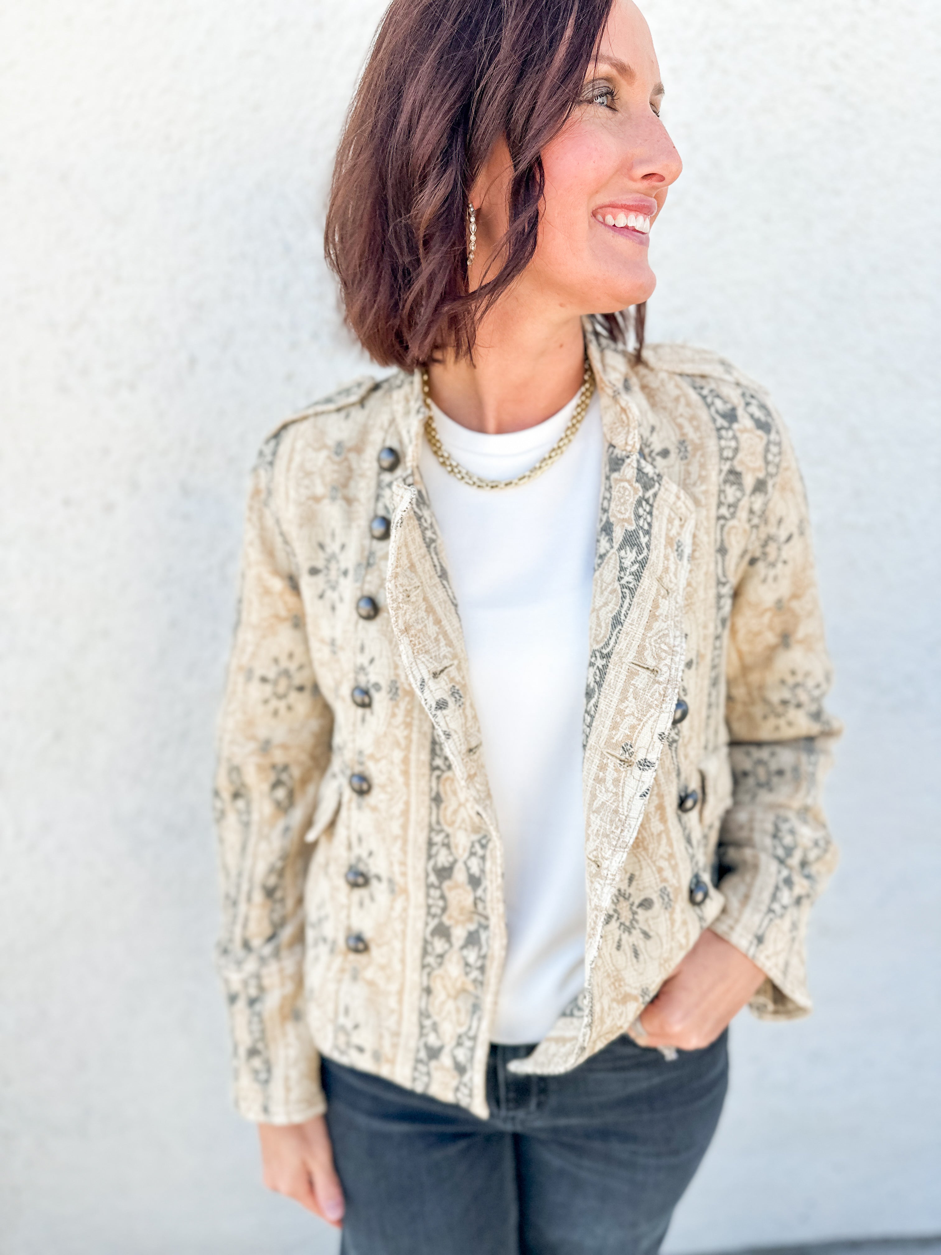 A woman wearing a beige and gray jacquard print jacket by Free People over a white t-shirt and dark gray jeans, standing against a light gray textured wall.