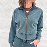A woman wearing a matching slate blue-gray funnel neck zip-up sweatshirt and drawstring barrel sweatpants, standing against a light textured wall.