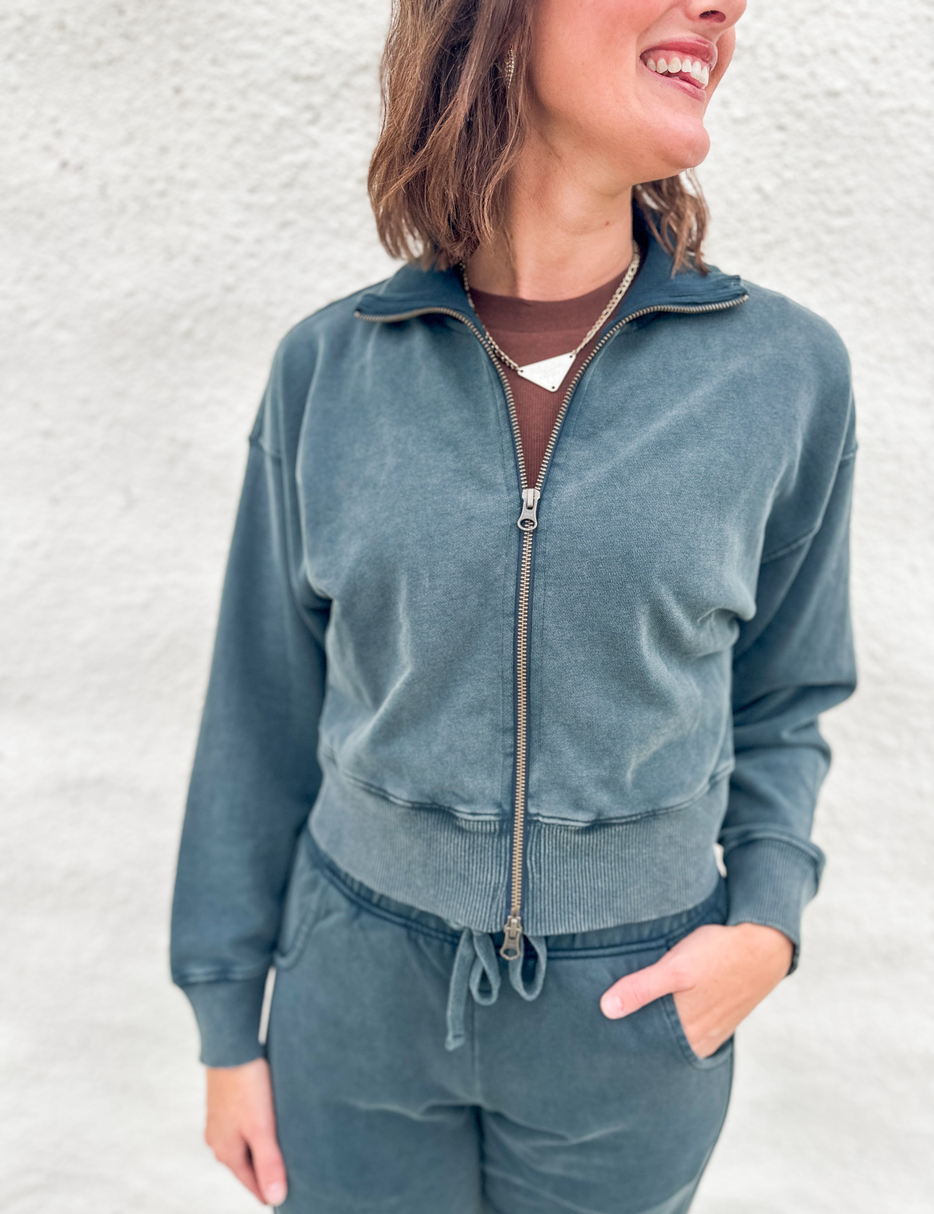 A woman wearing a matching slate blue-gray funnel neck zip-up sweatshirt and drawstring barrel sweatpants, standing against a light textured wall.