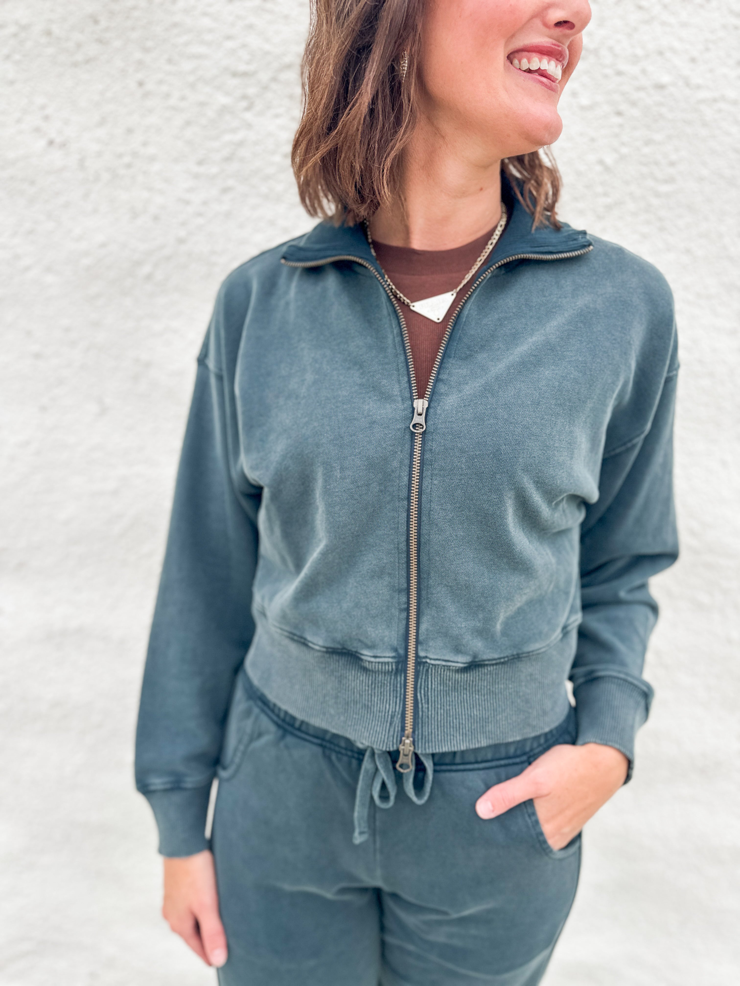 A woman wearing a matching slate blue-gray funnel neck zip-up sweatshirt and drawstring barrel sweatpants, standing against a light textured wall.