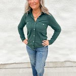 A woman with gray hair stands against a white stucco wall wearing a dark green long-sleeve button-up shirt, blue jeans, and tan boots.