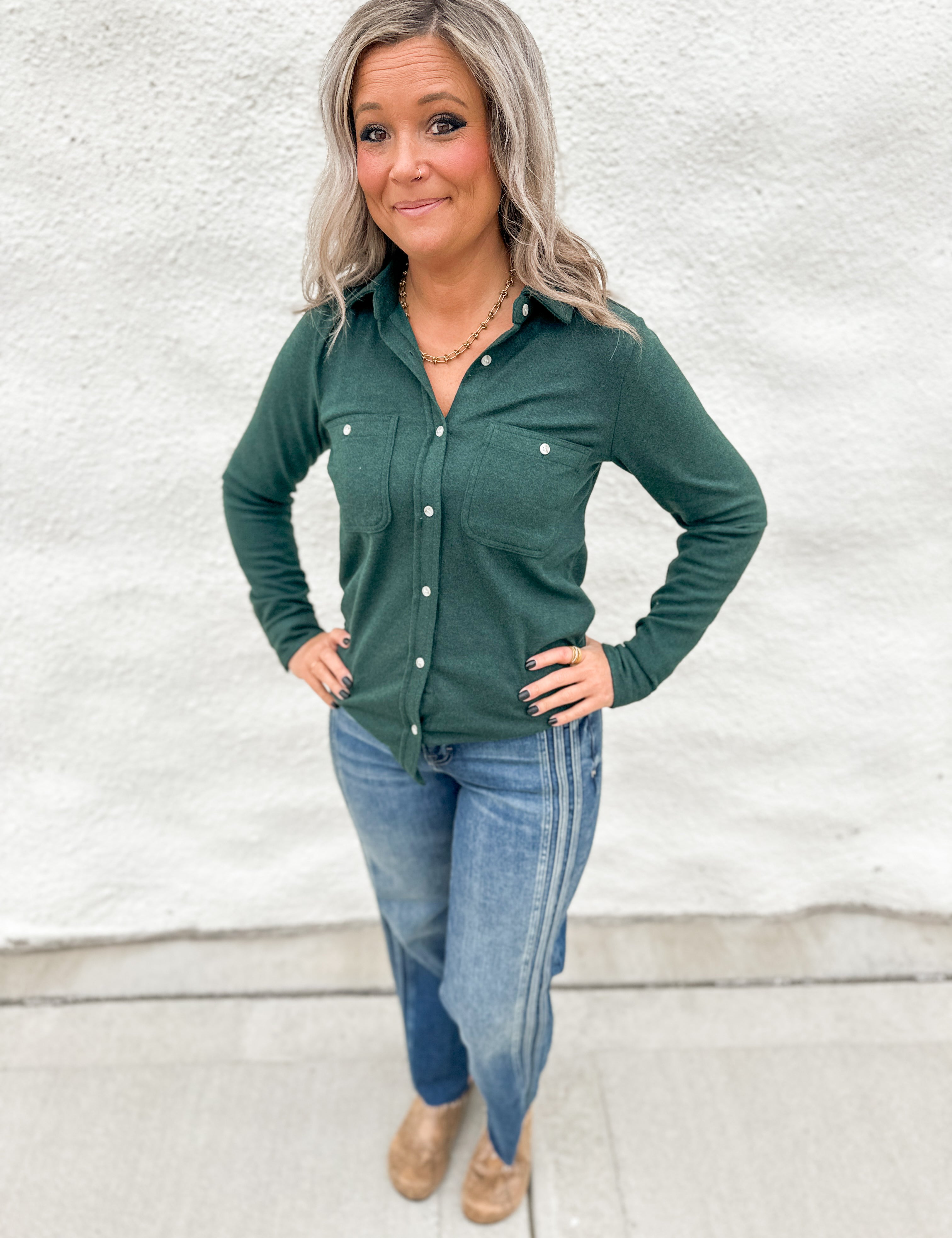 A woman with gray hair stands against a white stucco wall wearing a dark green long-sleeve button-up shirt, blue jeans, and tan boots.
