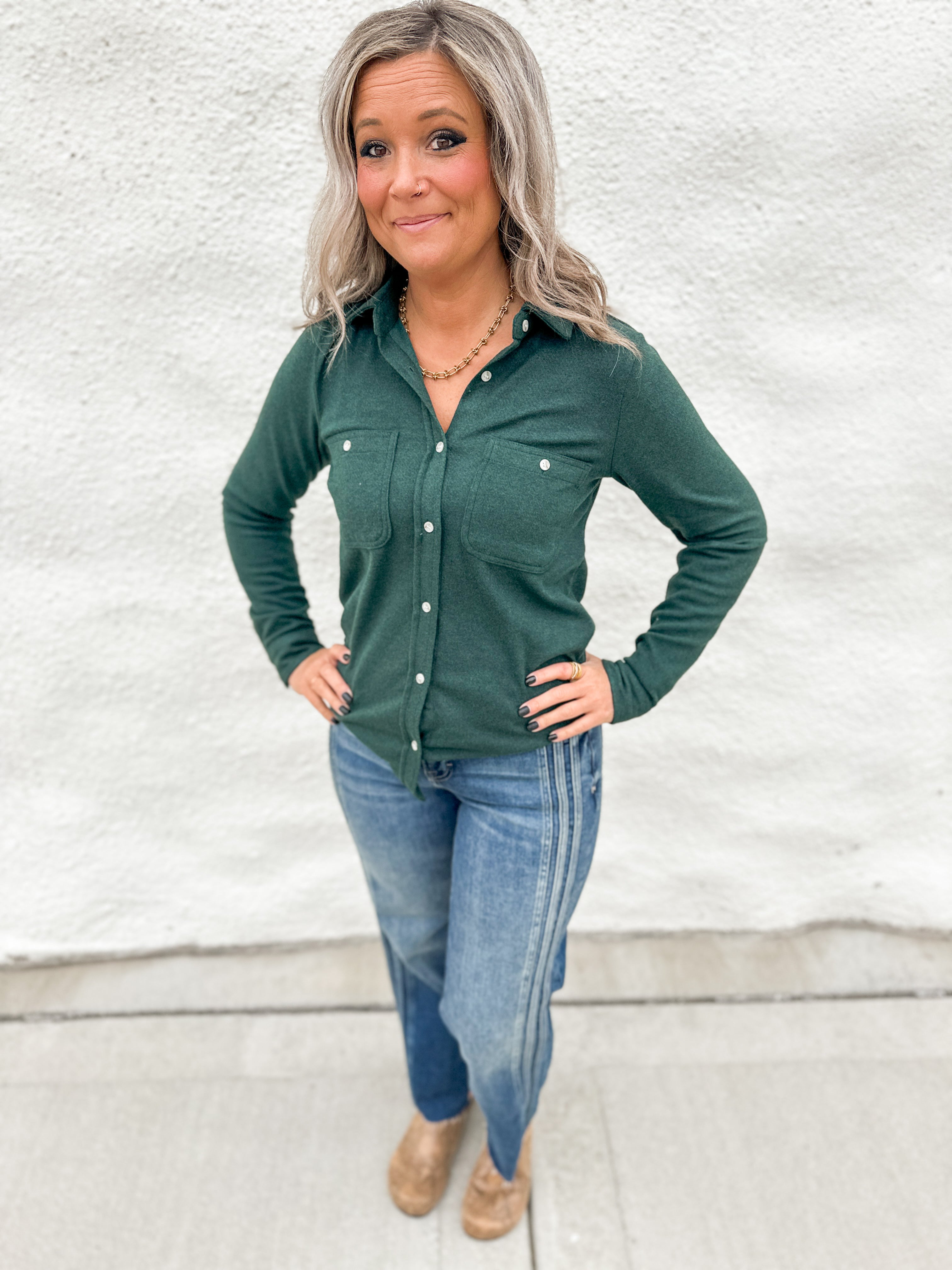 A woman with gray hair stands against a white stucco wall wearing a dark green long-sleeve button-up shirt, blue jeans, and tan boots.
