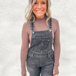 A woman wearing a fitted brown lightly ribbed long-sleeve mock neck top under washed black barrel overalls standing against a textured white wall