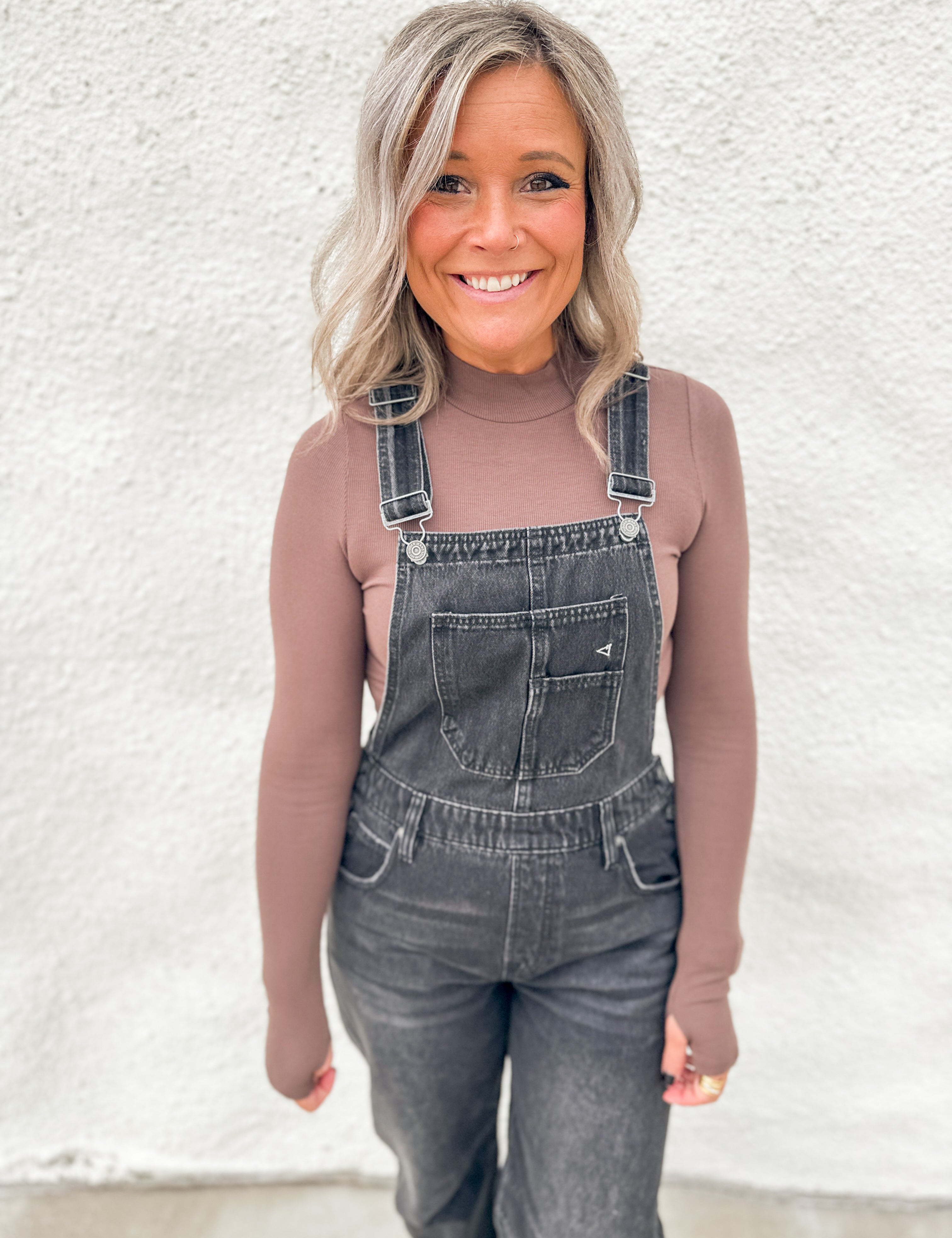 A woman wearing a fitted brown lightly ribbed long-sleeve mock neck top under washed black barrel overalls standing against a textured white wall