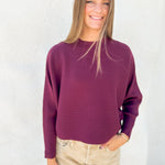 A woman wearing a ribbed long-sleeve crewneck sweater in deep burgundy, paired with beige pants, standing against a textured white wall.