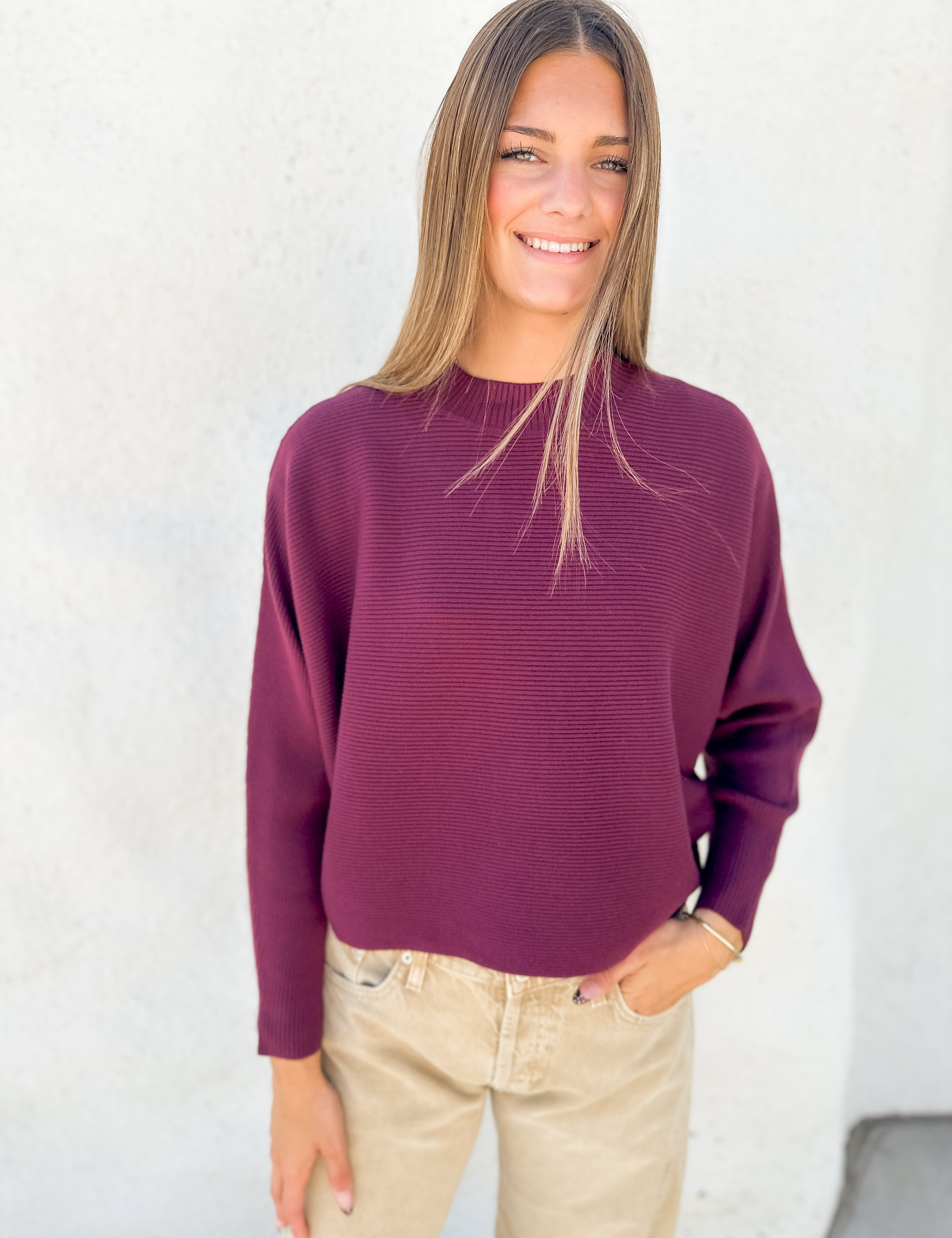 A woman wearing a ribbed long-sleeve crewneck sweater in deep burgundy, paired with beige pants, standing against a textured white wall.