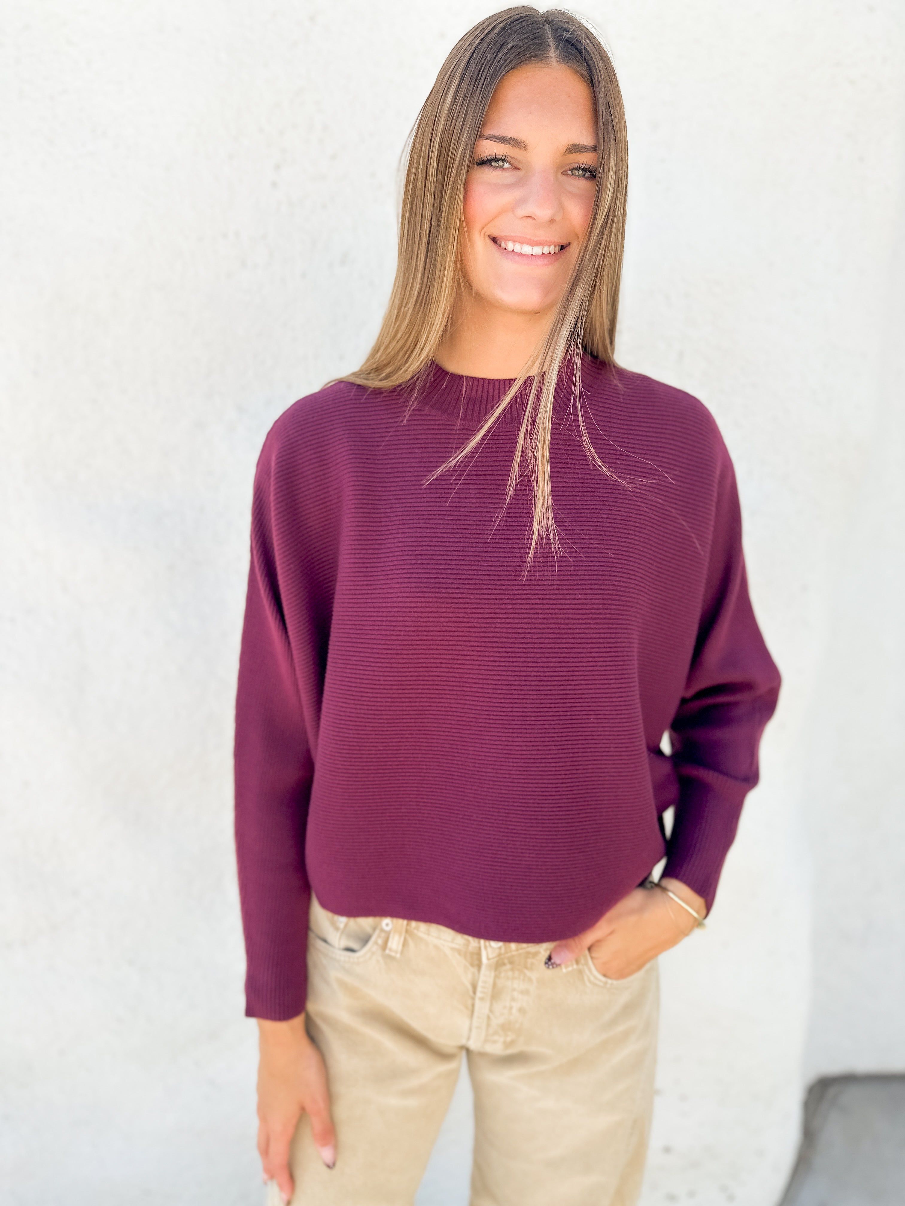 A woman wearing a ribbed long-sleeve crewneck sweater in deep burgundy, paired with beige pants, standing against a textured white wall.