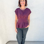 A woman modeling a short-sleeve peplum top in a deep plum purple color, paired with dark gray pants and black heels, standing against a textured white stucco wall.