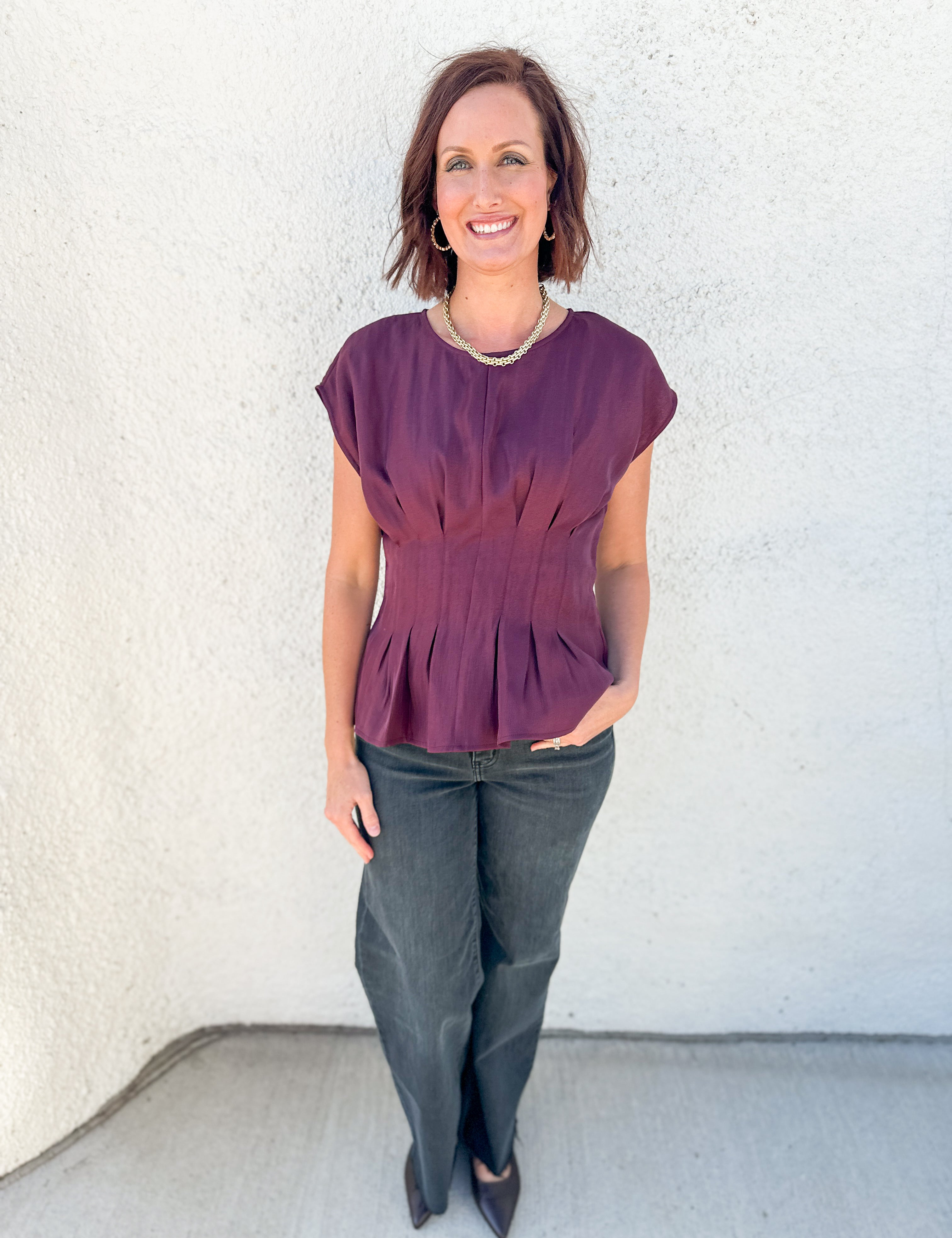 A woman modeling a short-sleeve peplum top in a deep plum purple color, paired with dark gray pants and black heels, standing against a textured white stucco wall.