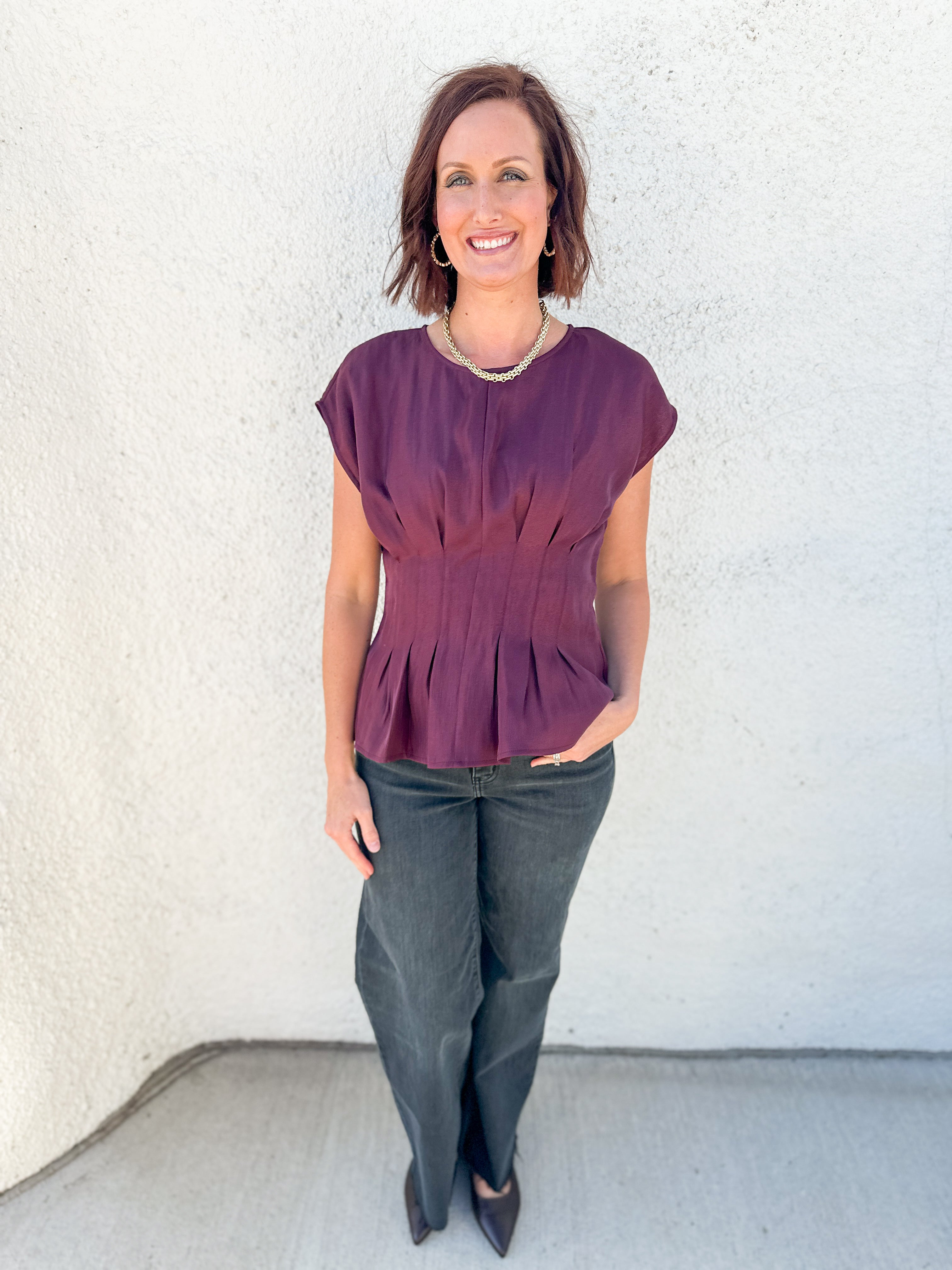 A woman modeling a short-sleeve peplum top in a deep plum purple color, paired with dark gray pants and black heels, standing against a textured white stucco wall.