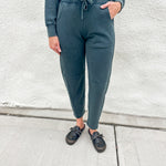 A person wearing dark washed teal barrel jogger sweatpants with a drawstring waist, paired with a matching funnel neck zip up and black loafer clog slip-on shoes, standing against a light gray textured wall.