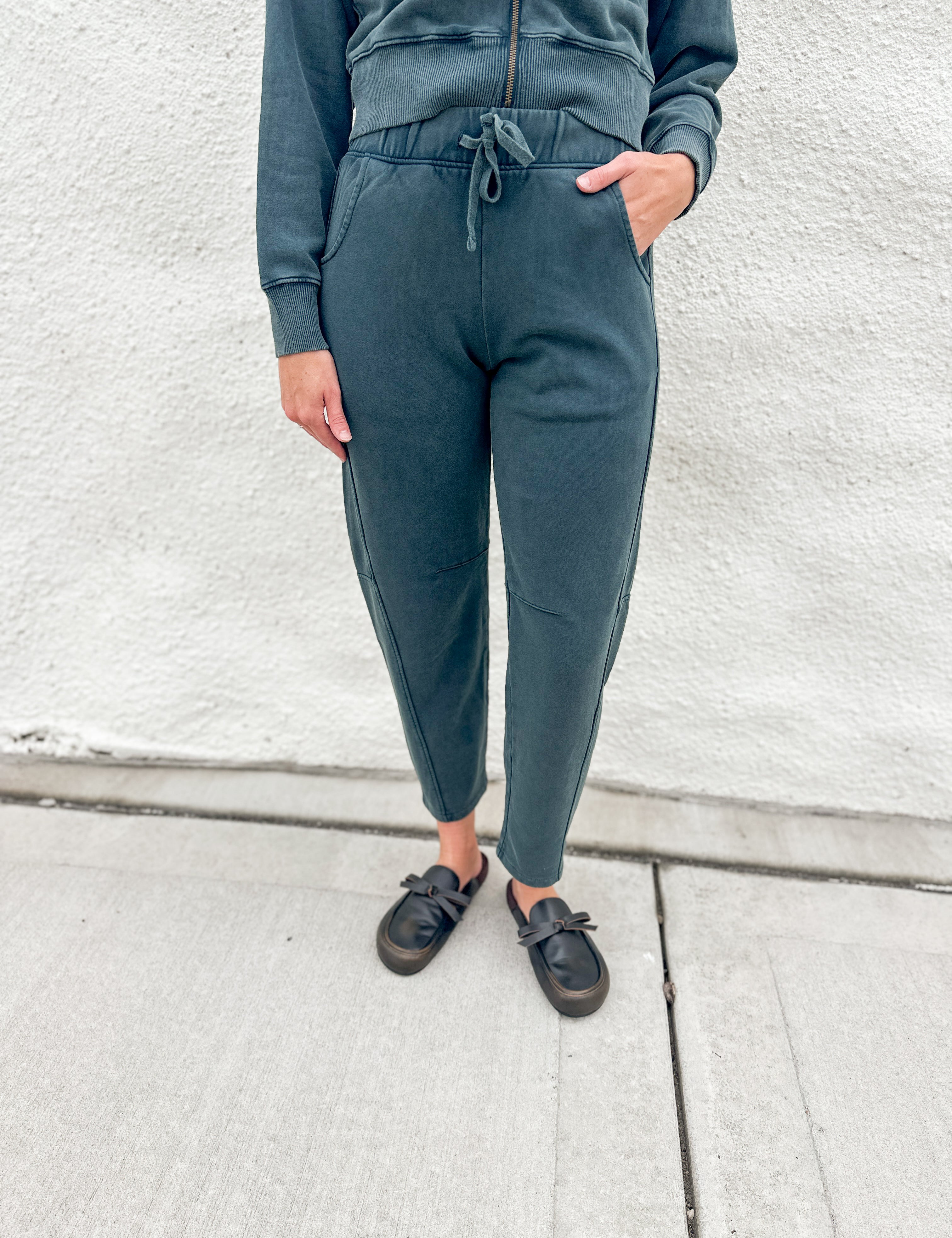 A person wearing dark washed teal barrel jogger sweatpants with a drawstring waist, paired with a matching funnel neck zip up and black loafer clog slip-on shoes, standing against a light gray textured wall.