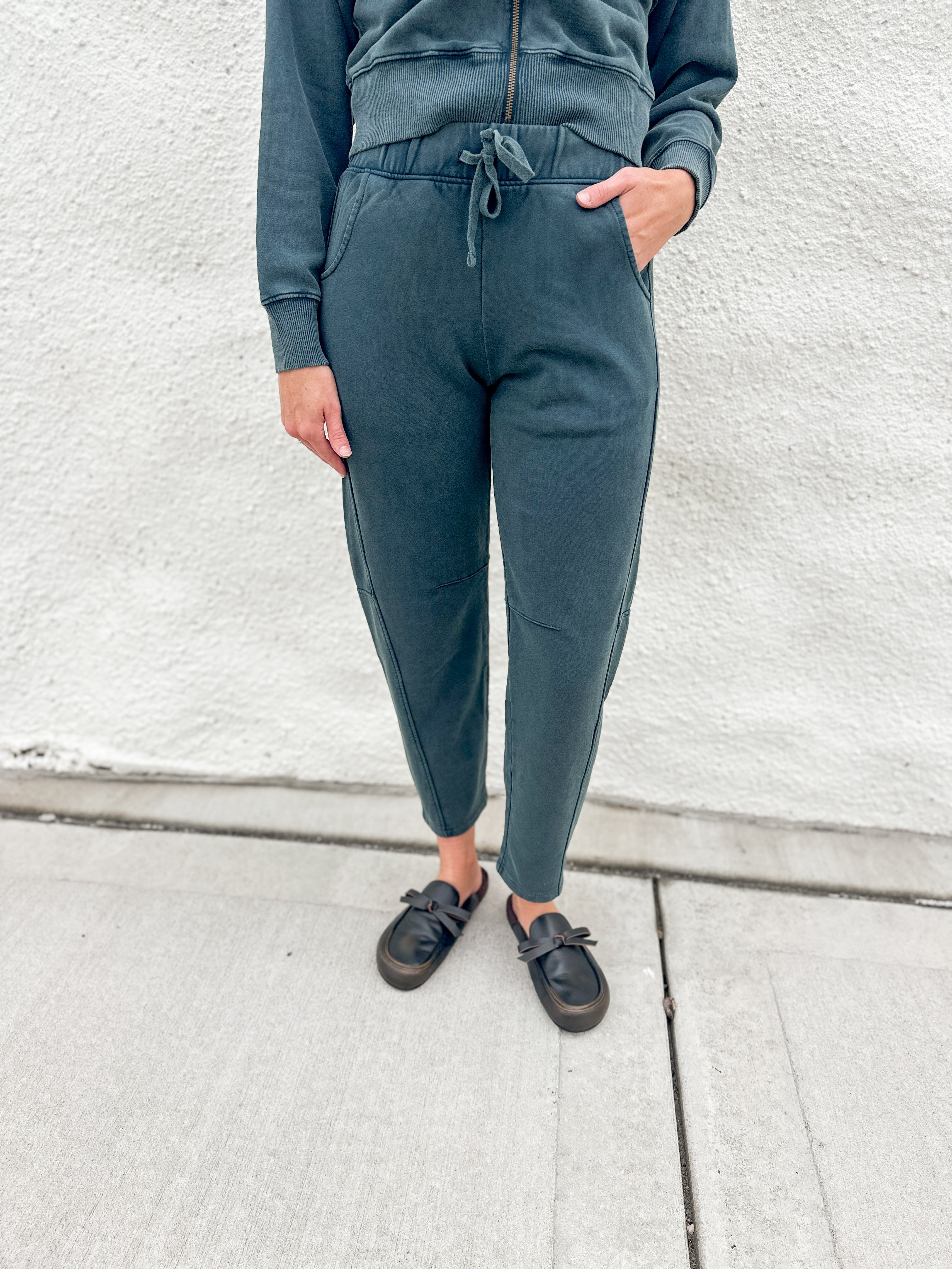A person wearing dark washed teal barrel jogger sweatpants with a drawstring waist, paired with a matching funnel neck zip up and black loafer clog slip-on shoes, standing against a light gray textured wall.