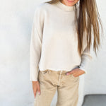 A woman wearing a cream-colored knit mock neck sweater and beige pants, standing against a textured white wall.