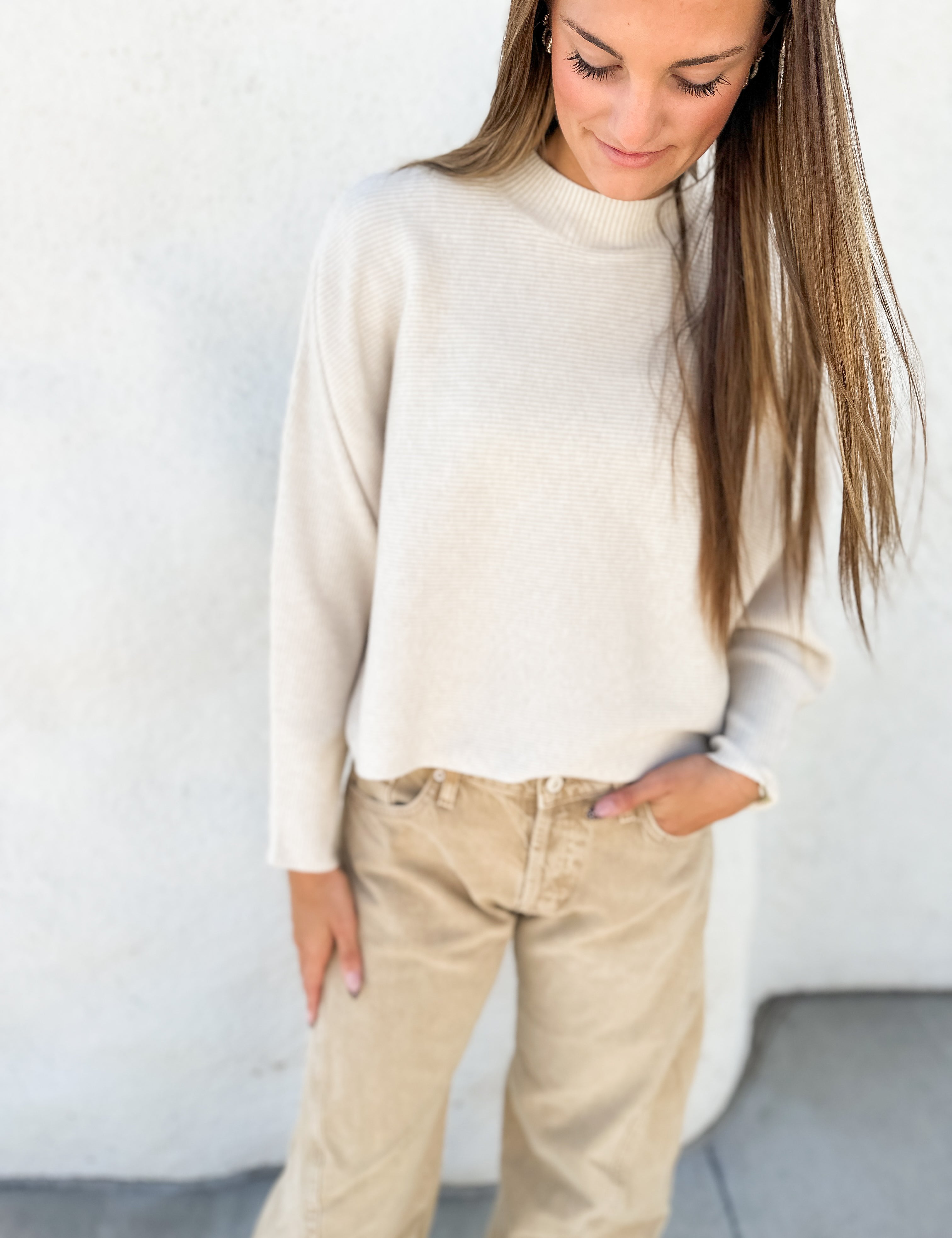 A woman wearing a cream-colored knit mock neck sweater and beige pants, standing against a textured white wall.