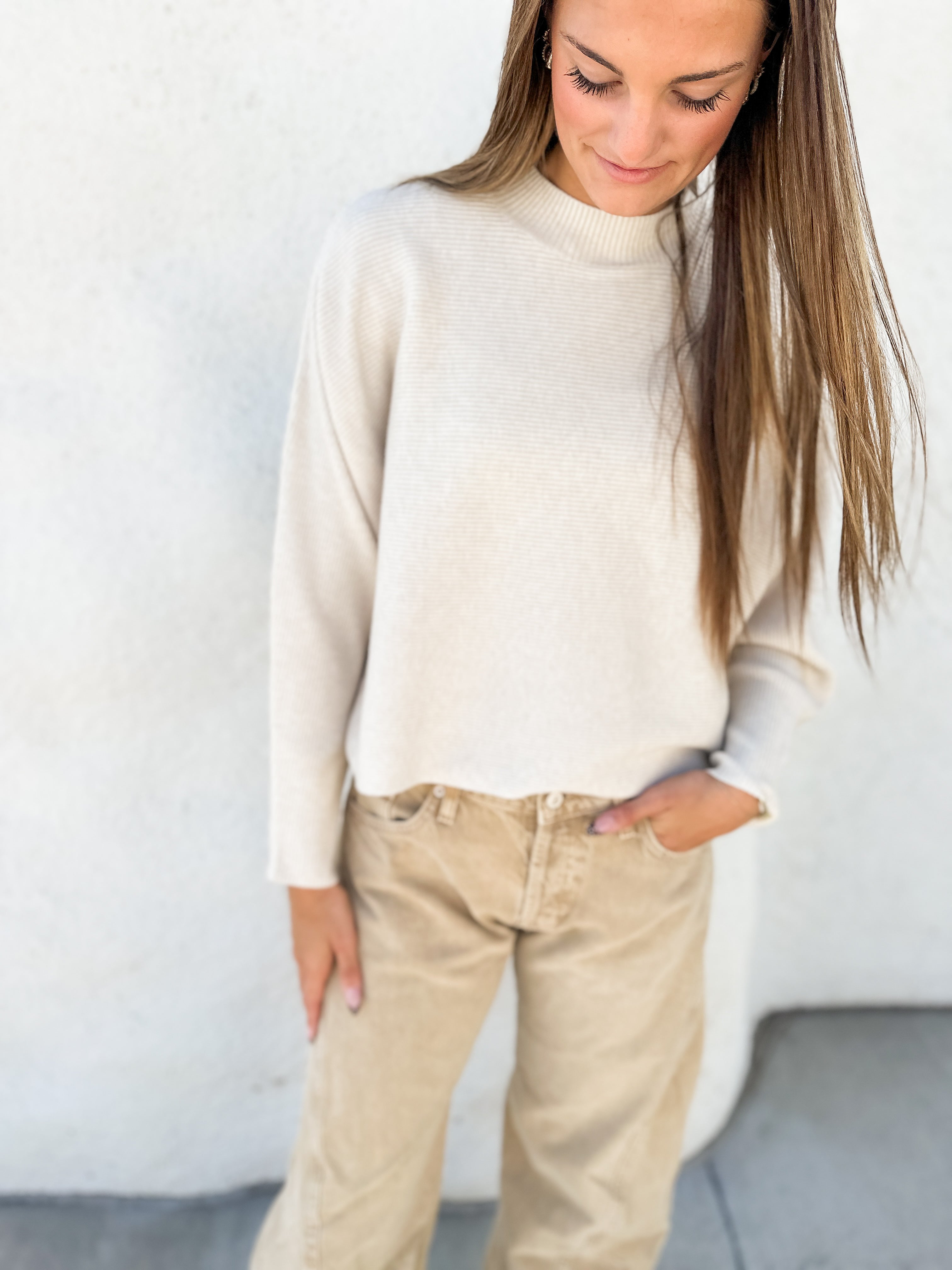 A woman wearing a cream-colored knit mock neck sweater and beige pants, standing against a textured white wall.