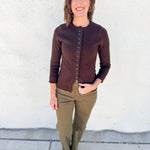 A woman stands outdoors against a white stucco wall wearing a brown button-front cardigan, olive green pants, and black shoes.