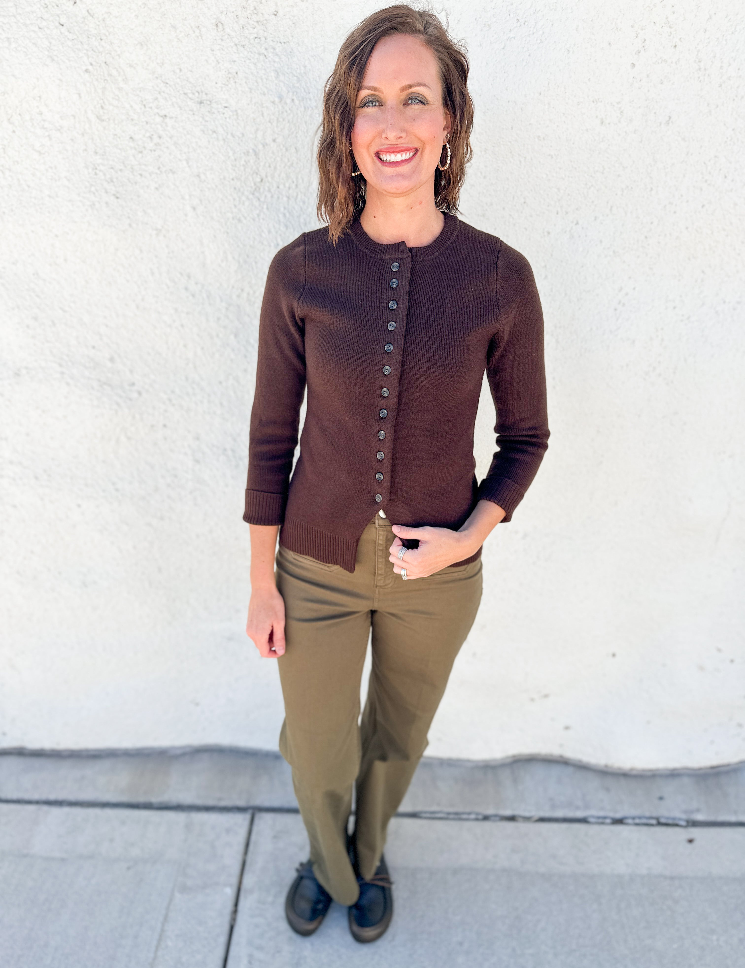A woman stands outdoors against a white stucco wall wearing a brown button-front cardigan, olive green pants, and black shoes.