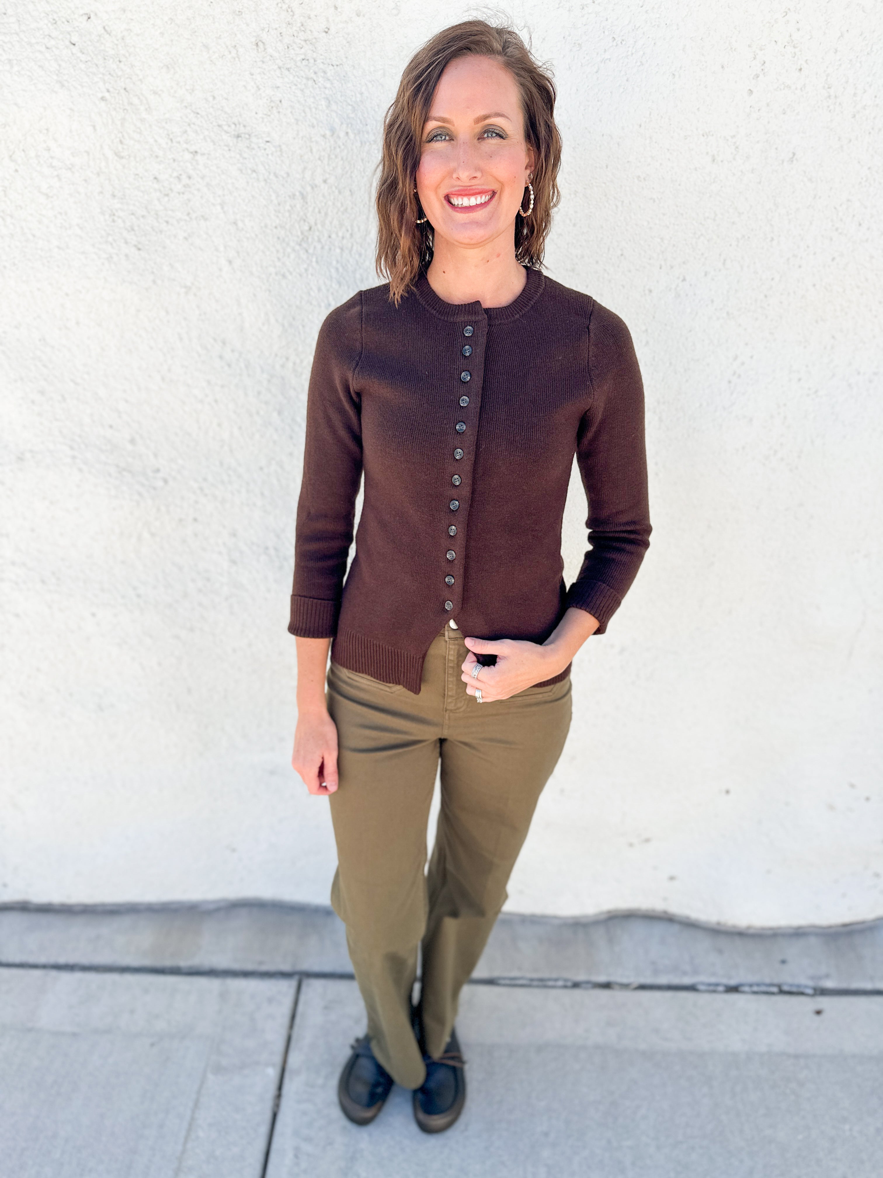 A woman stands outdoors against a white stucco wall wearing a brown button-front cardigan, olive green pants, and black shoes.