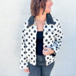 A woman wearing a white jacket with black polka dots and a black faux leather collar, styled with a black top and dark gray jeans, standing against a textured white wall.