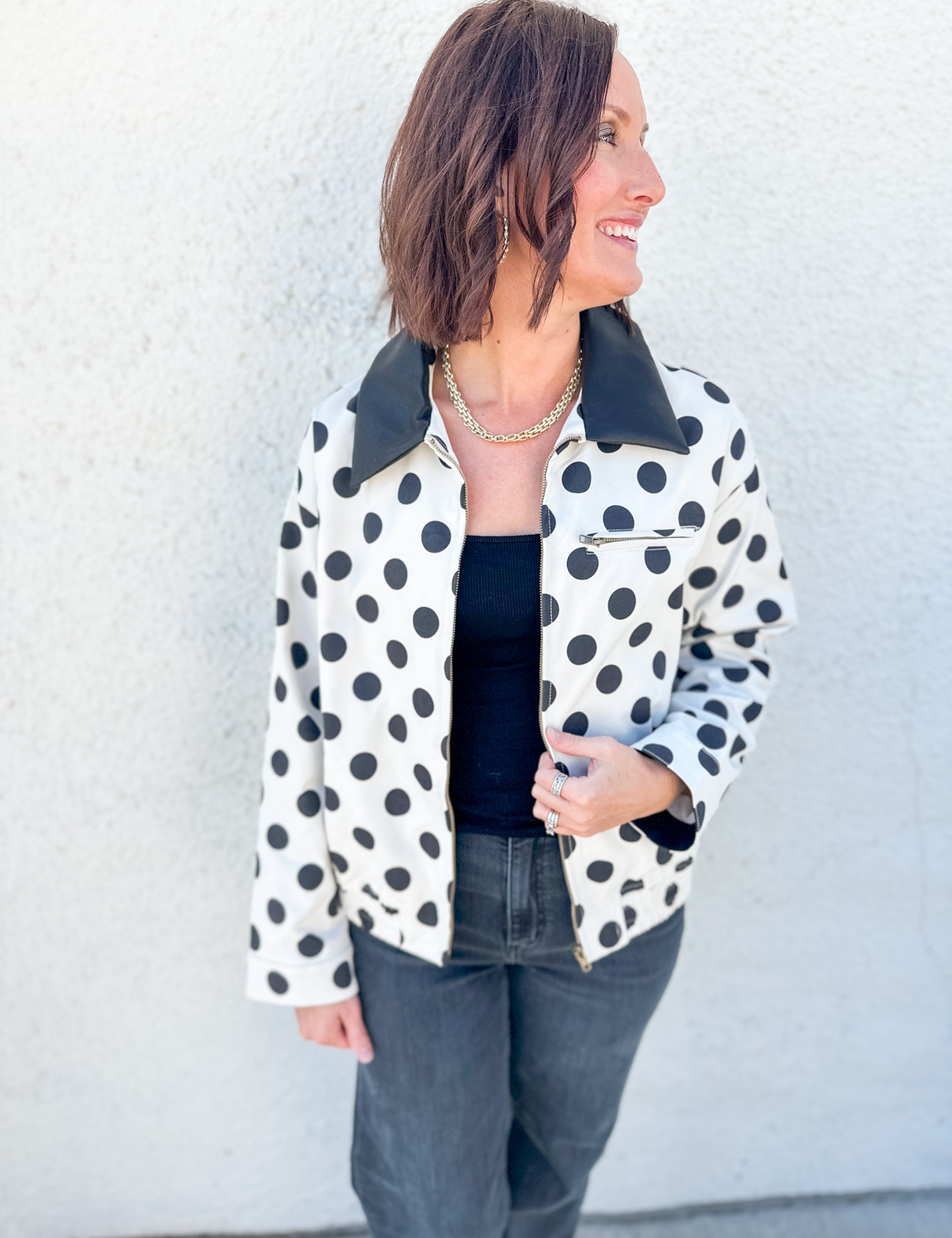 A woman wearing a white jacket with black polka dots and a black faux leather collar, styled with a black top and dark gray jeans, standing against a textured white wall.