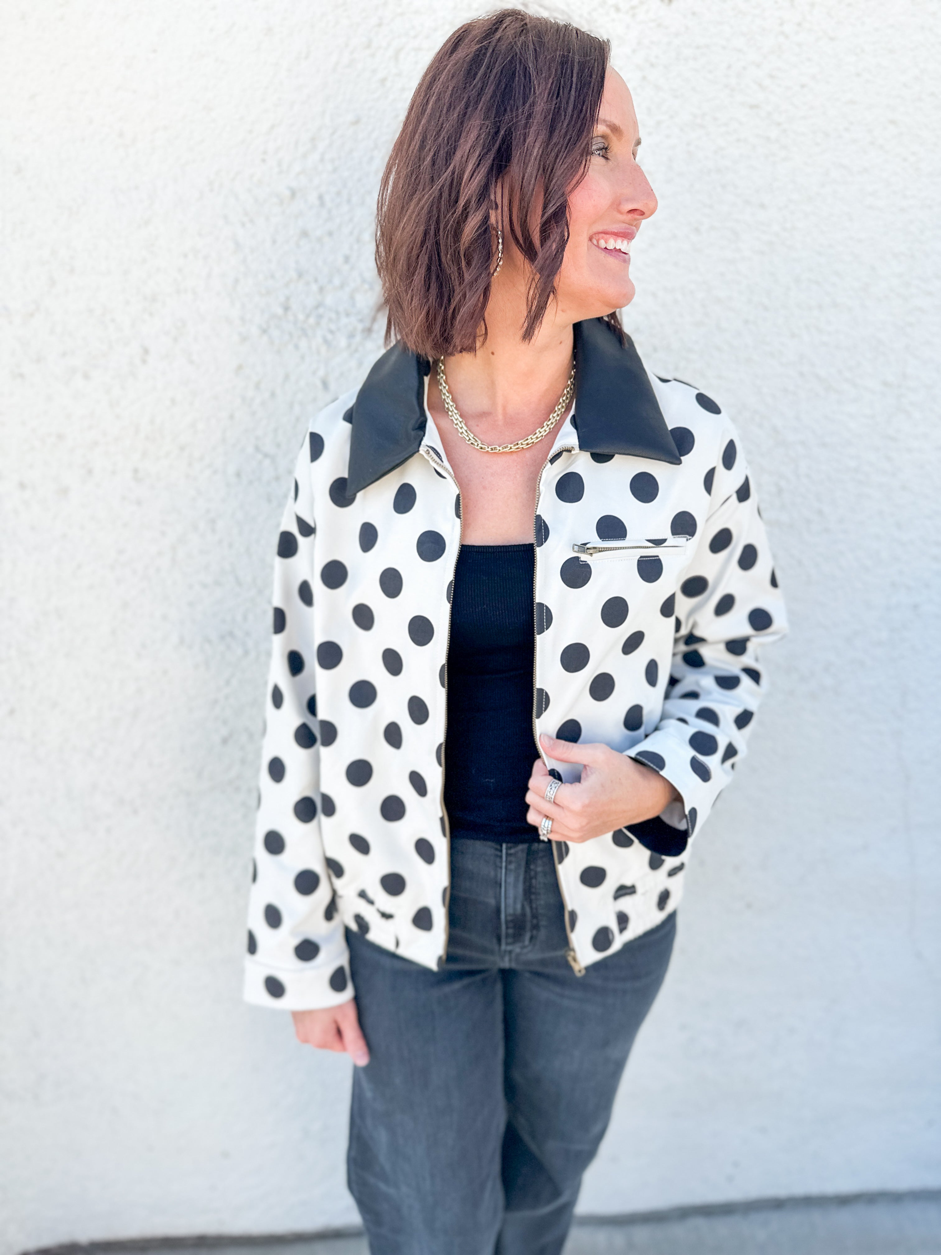 A woman wearing a white jacket with black polka dots and a black faux leather collar, styled with a black top and dark gray jeans, standing against a textured white wall.