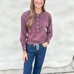 A woman modeling a deep purple long-sleeve henley top with silver buttons, paired with dark blue jeans, standing against a textured white wall.