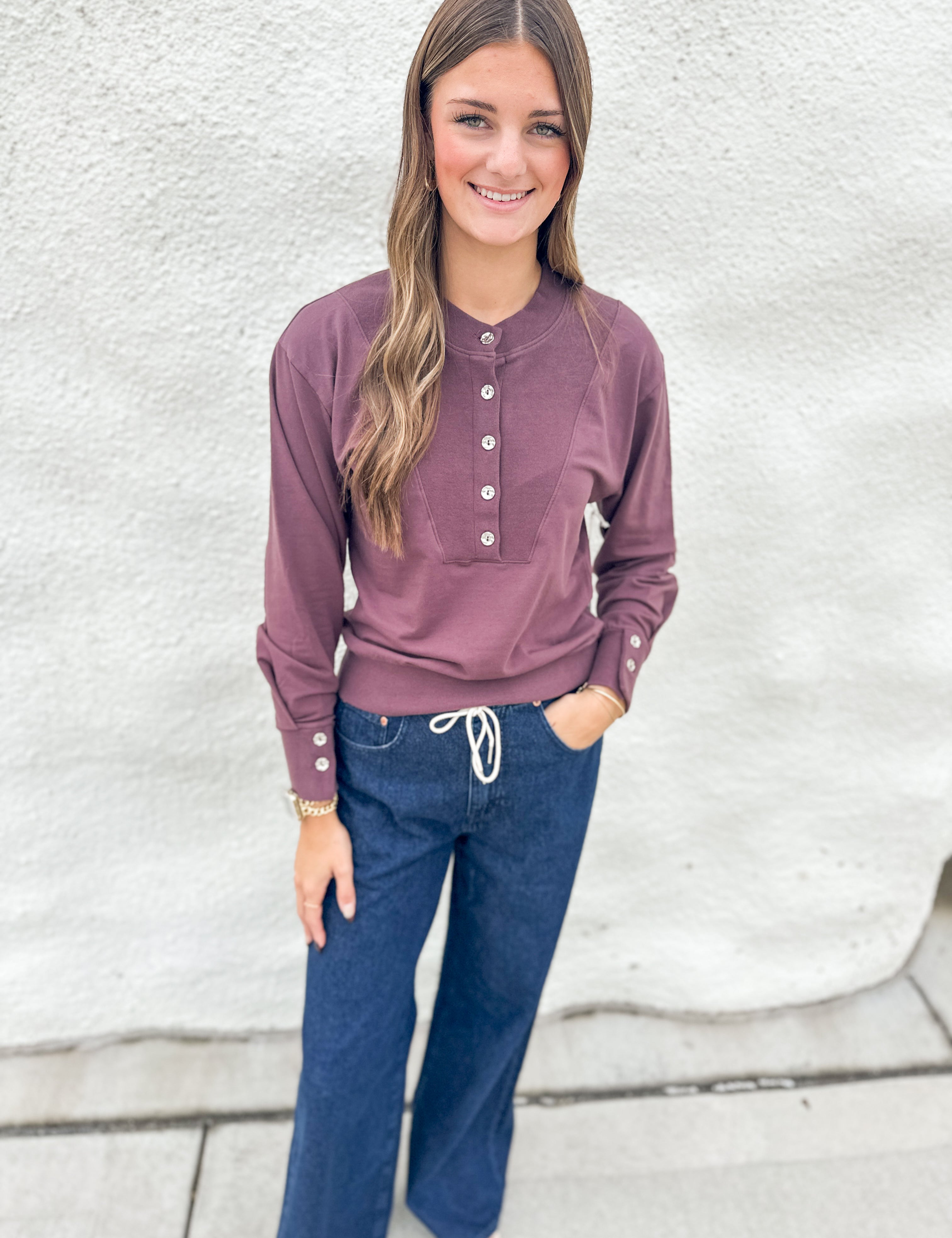 A woman modeling a deep purple long-sleeve henley top with silver buttons, paired with dark blue jeans, standing against a textured white wall.