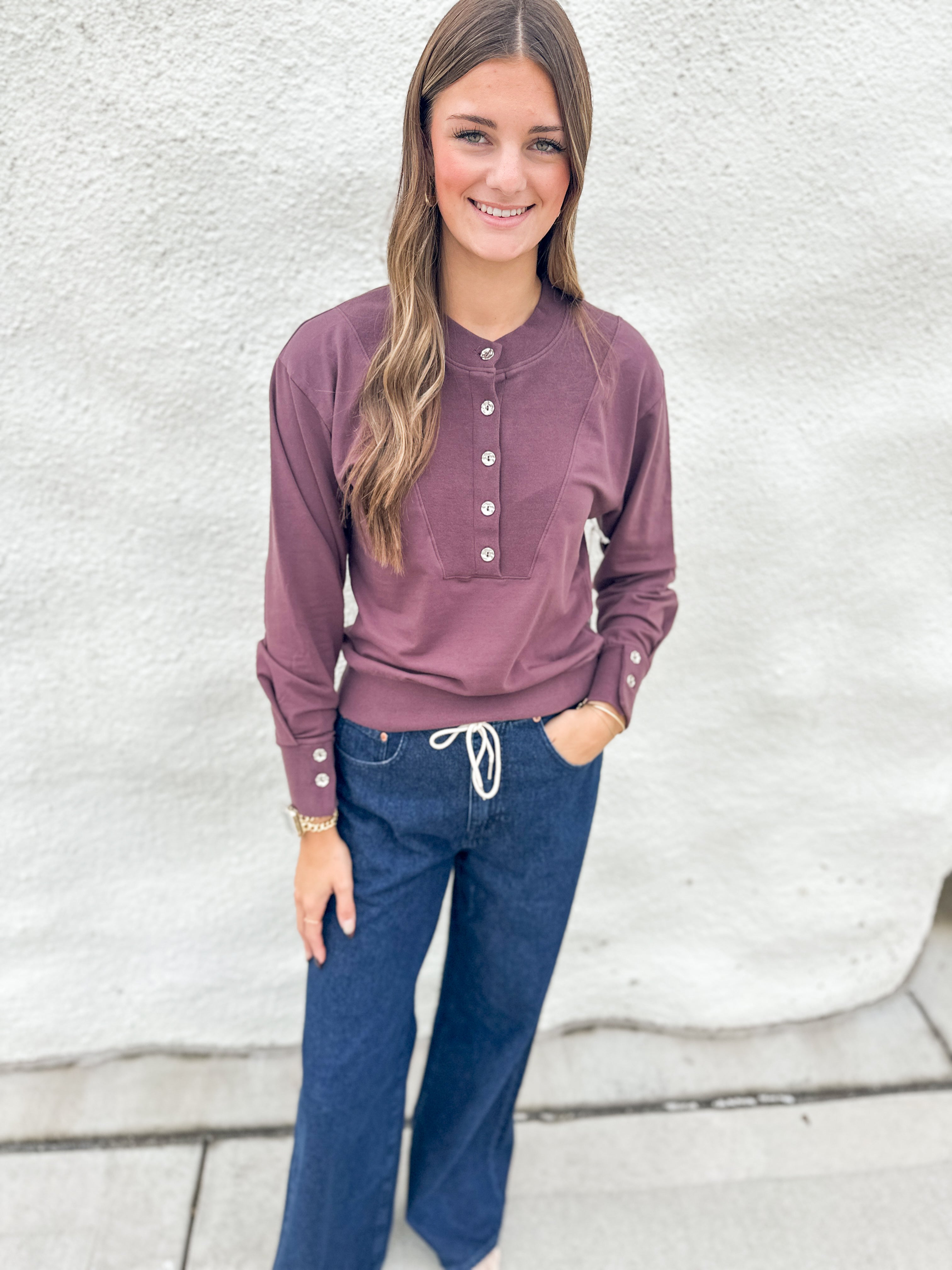A woman modeling a deep purple long-sleeve henley top with silver buttons, paired with dark blue jeans, standing against a textured white wall.