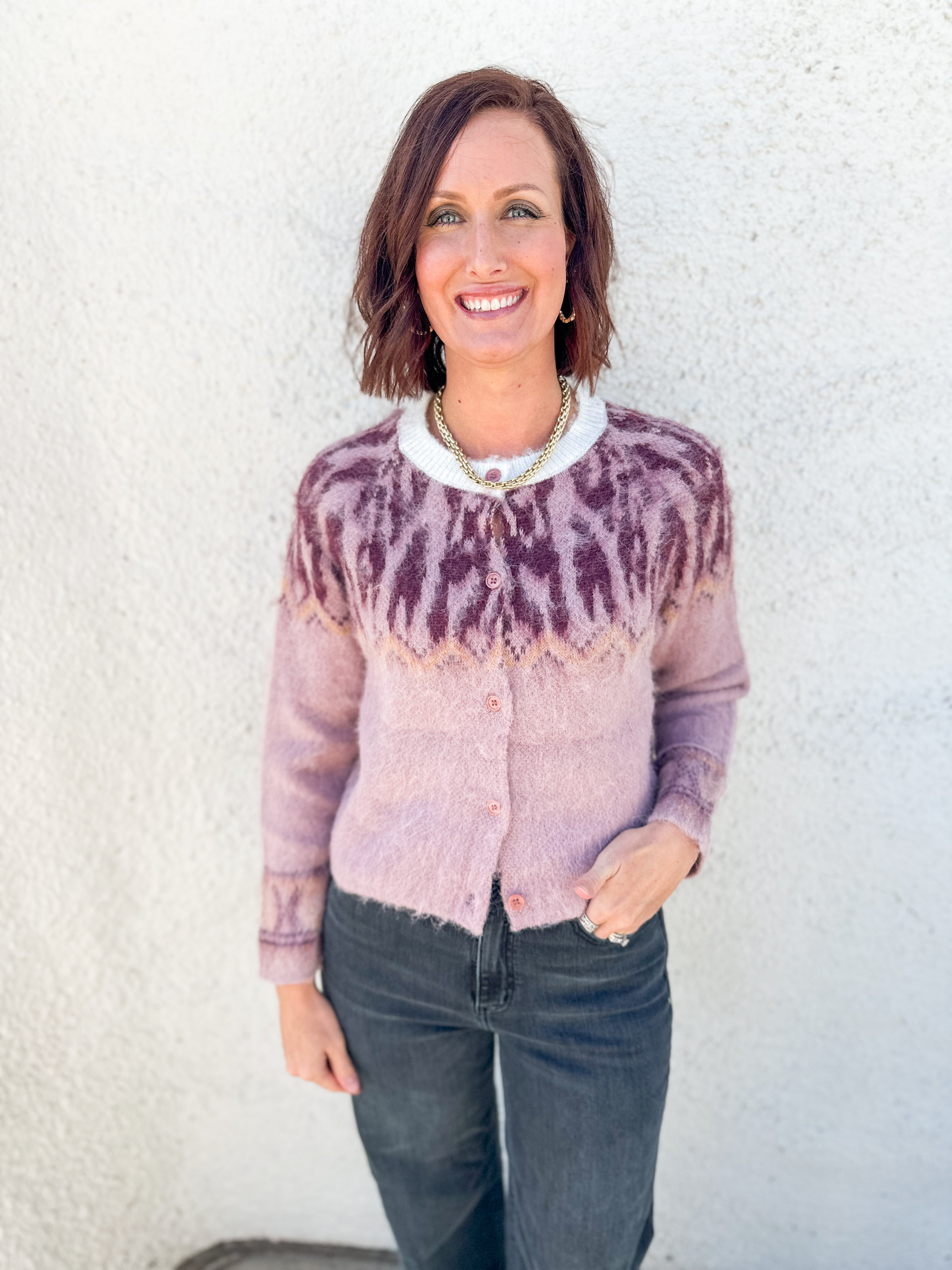 A smiling woman with short brown hair stands against a white textured wall wearing a fuzzy knit cardigan with a maroon and dusty pink Fair Isle yoke pattern, paired with dark gray jeans.