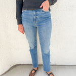 A person wearing light-wash blue jeans, a dark charcoal ribbed sweater, and brown Mary Jane flats, standing against a textured off-white wall.