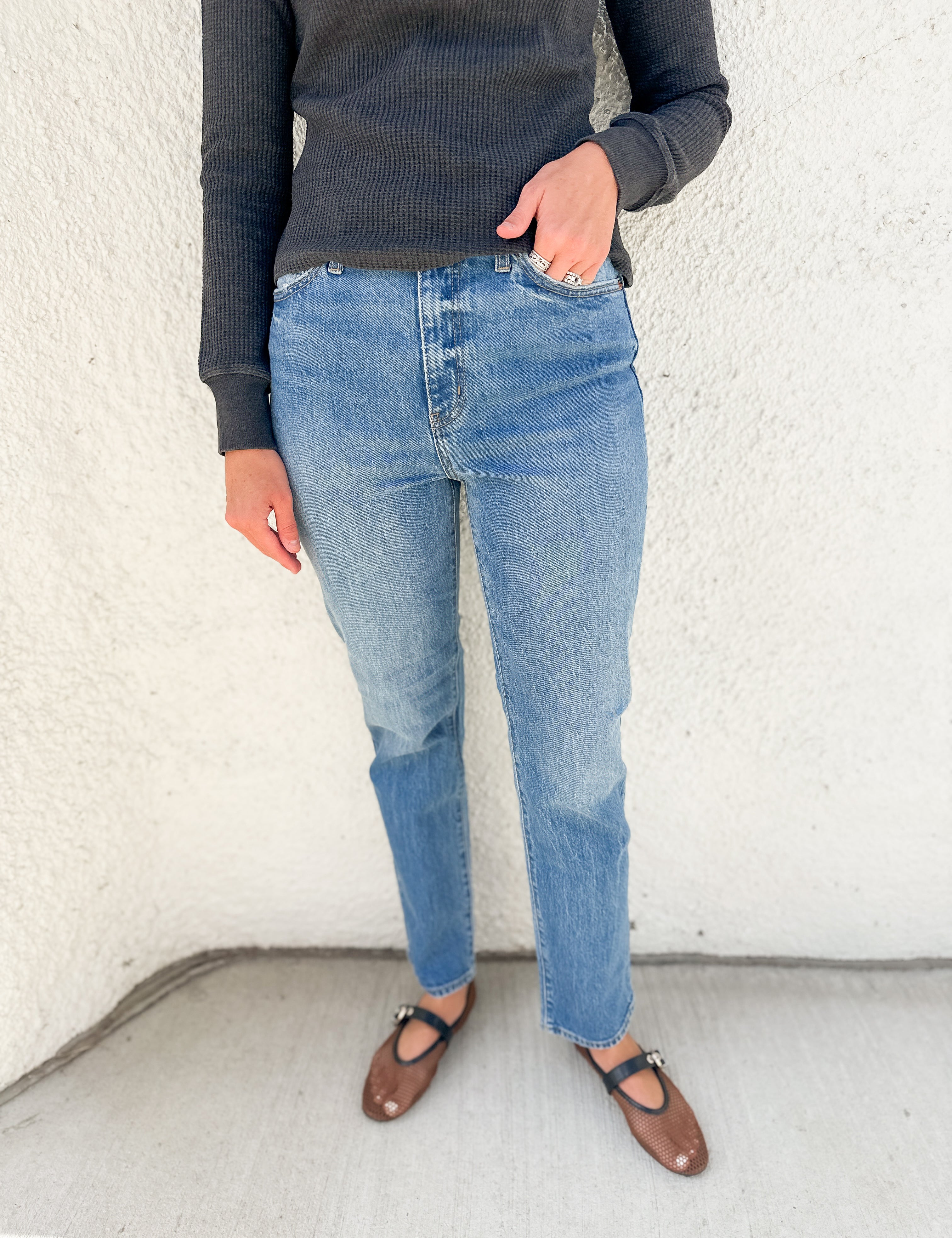 A person wearing light-wash blue jeans, a dark charcoal ribbed sweater, and brown Mary Jane flats, standing against a textured off-white wall.