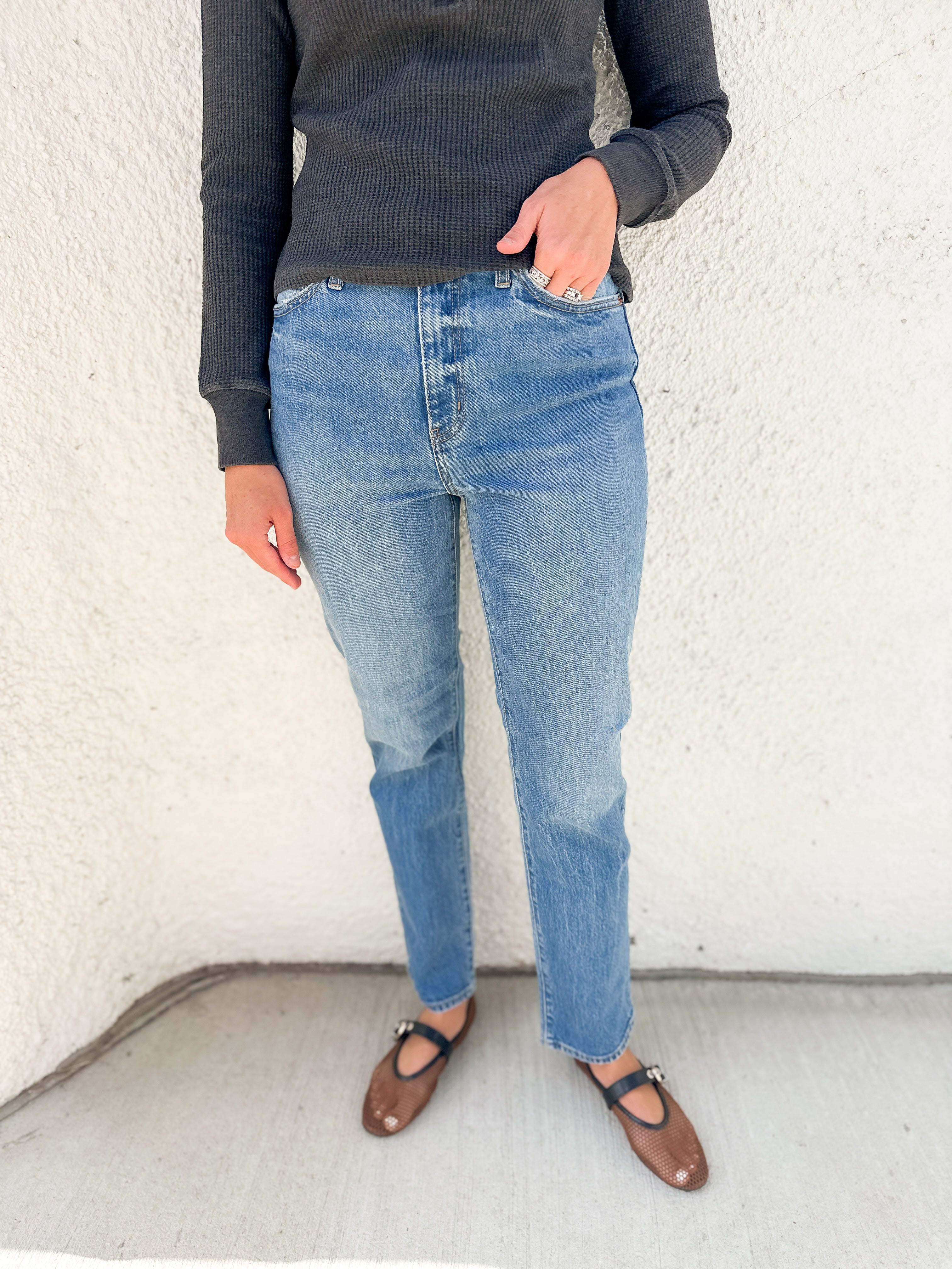A person wearing light-wash blue jeans, a dark charcoal ribbed sweater, and brown Mary Jane flats, standing against a textured off-white wall.