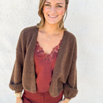 A woman wearing a brown fuzzy knit cardigan over a rust-red lace-trim camisole and matching rust-red pants, standing against a white stucco wall.
