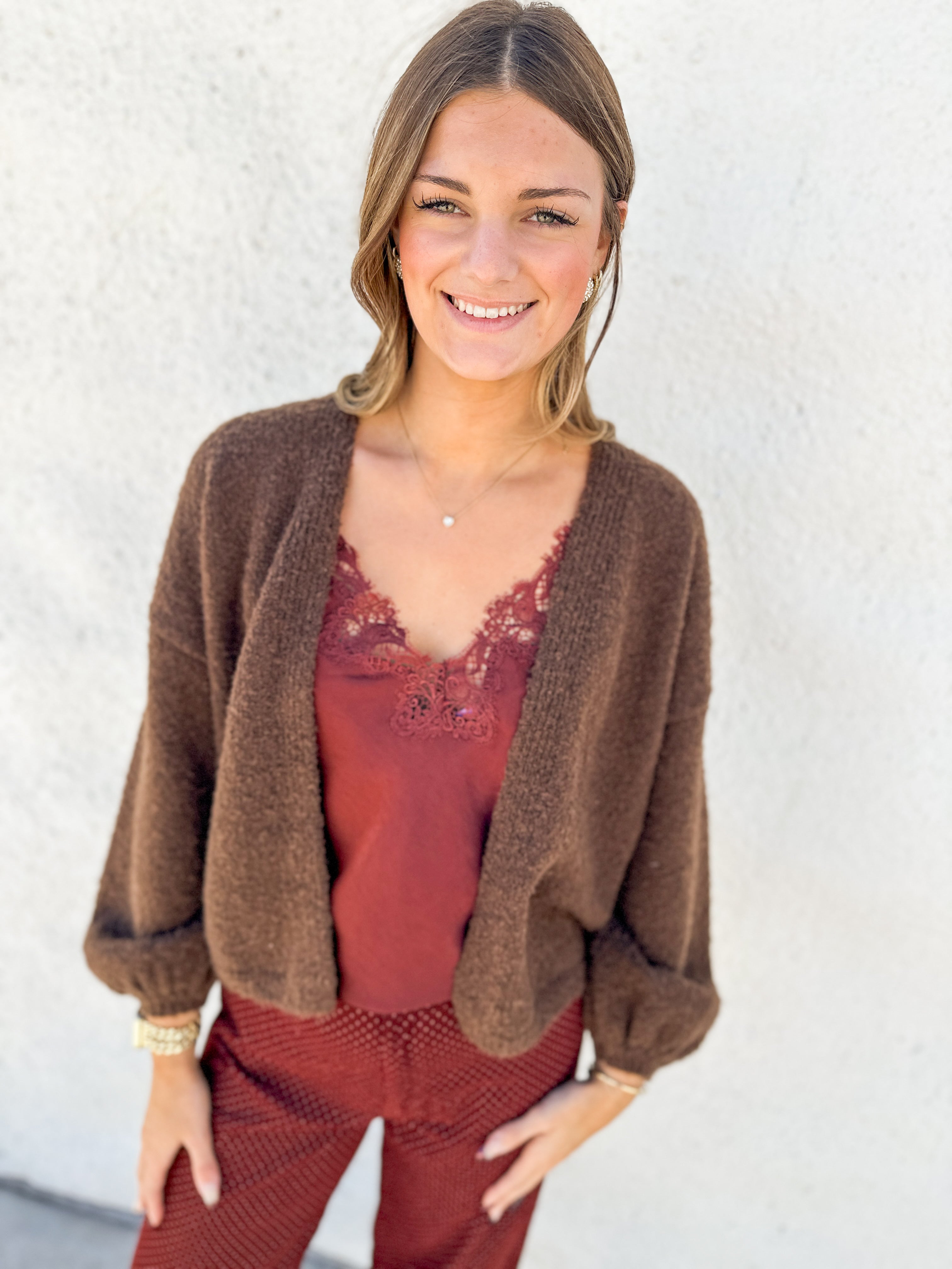 A woman wearing a brown fuzzy knit cardigan over a rust-red lace-trim camisole and matching rust-red pants, standing against a white stucco wall.