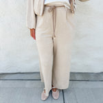 A person wearing a beige wide-leg drawstring pantsuit with suede accent belt with a white top and beige flats, standing against a textured off-white wall.