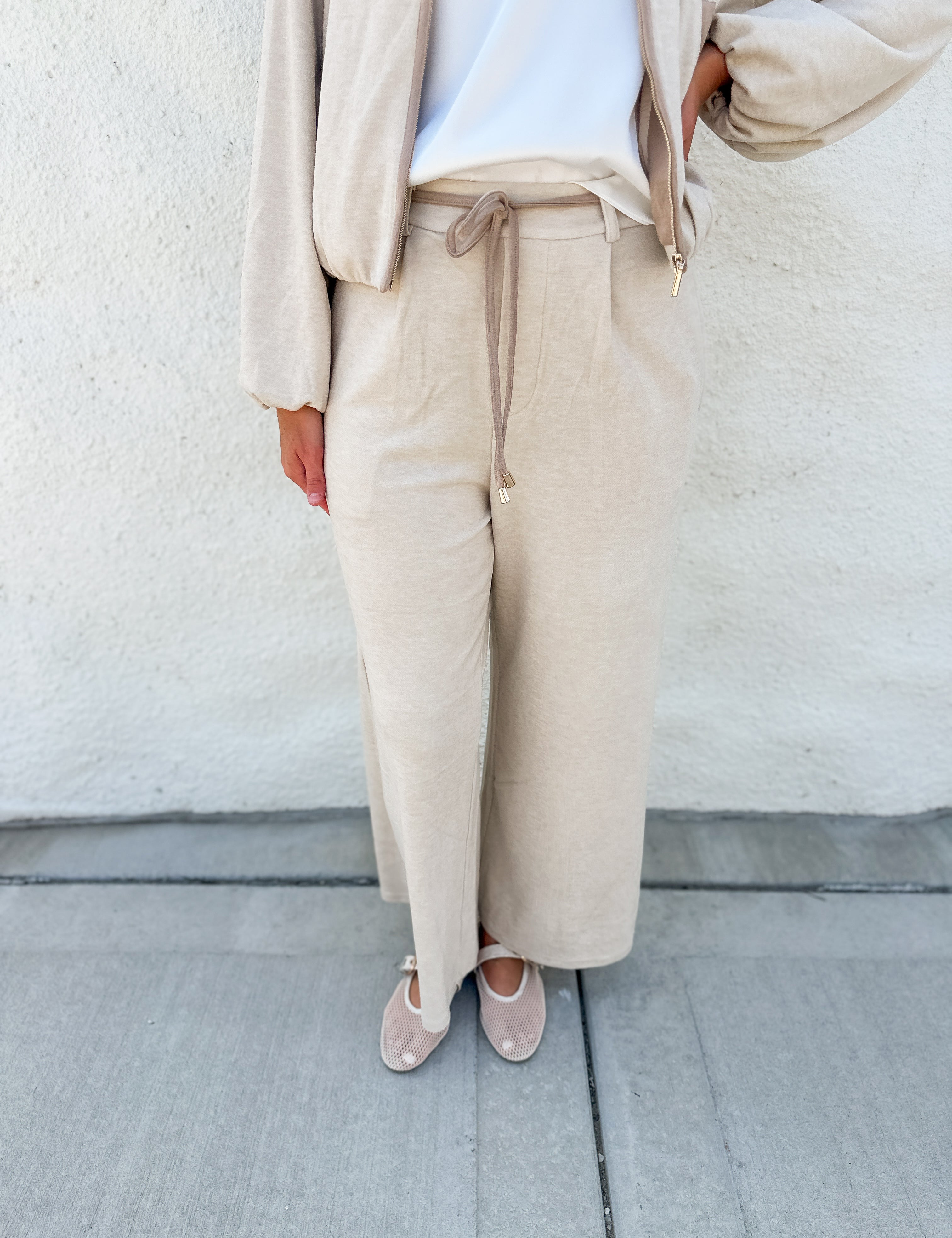 A person wearing a beige wide-leg drawstring pantsuit with suede accent belt with a white top and beige flats, standing against a textured off-white wall.