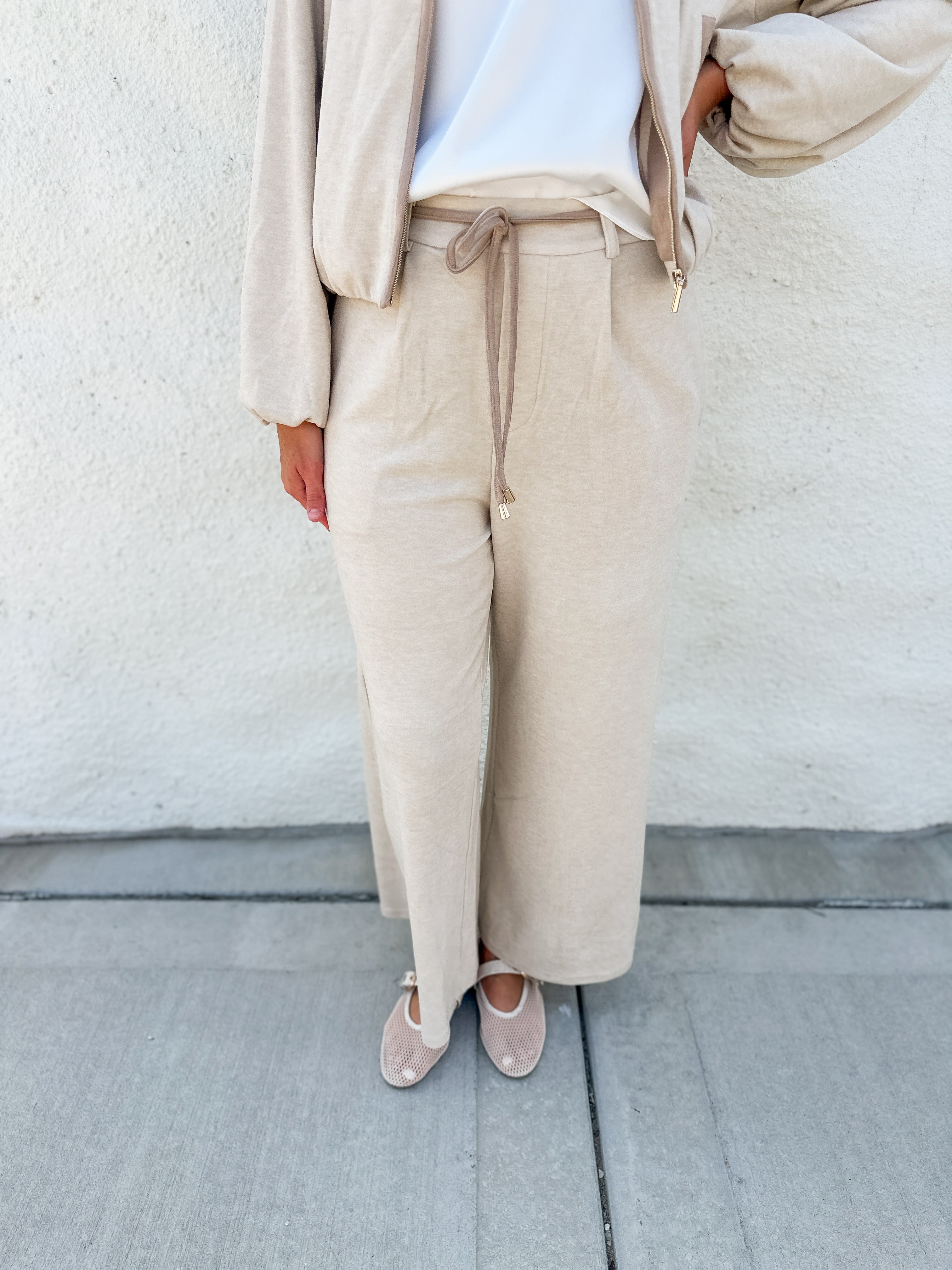 A person wearing a beige wide-leg drawstring pantsuit with suede accent belt with a white top and beige flats, standing against a textured off-white wall.