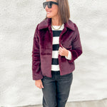 A woman wearing a burgundy faux fur jacket over a black and white striped sweater, dark jeans, and black boots, standing against a white textured wall.