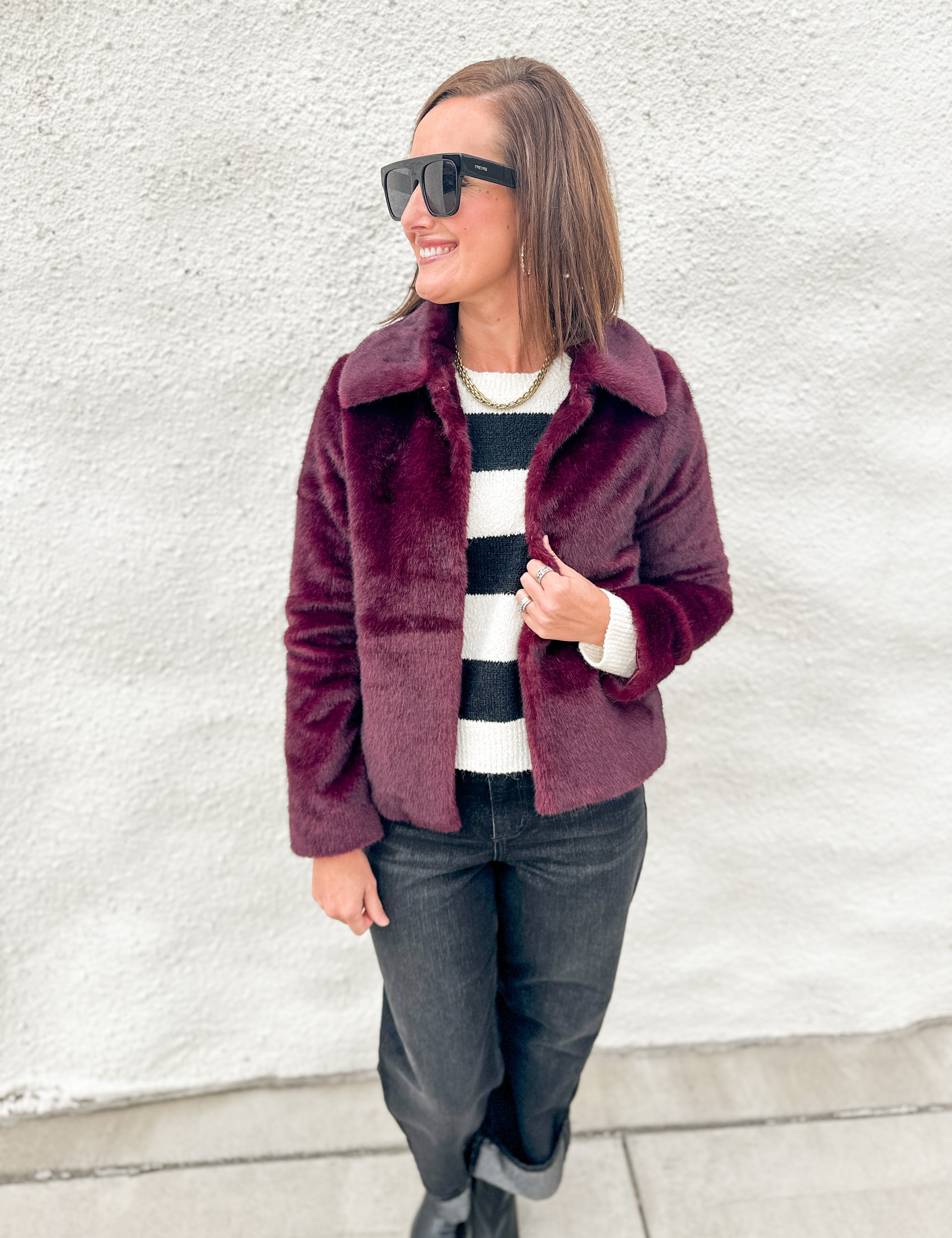 A woman wearing a burgundy faux fur jacket over a black and white striped sweater, dark jeans, and black boots, standing against a white textured wall.