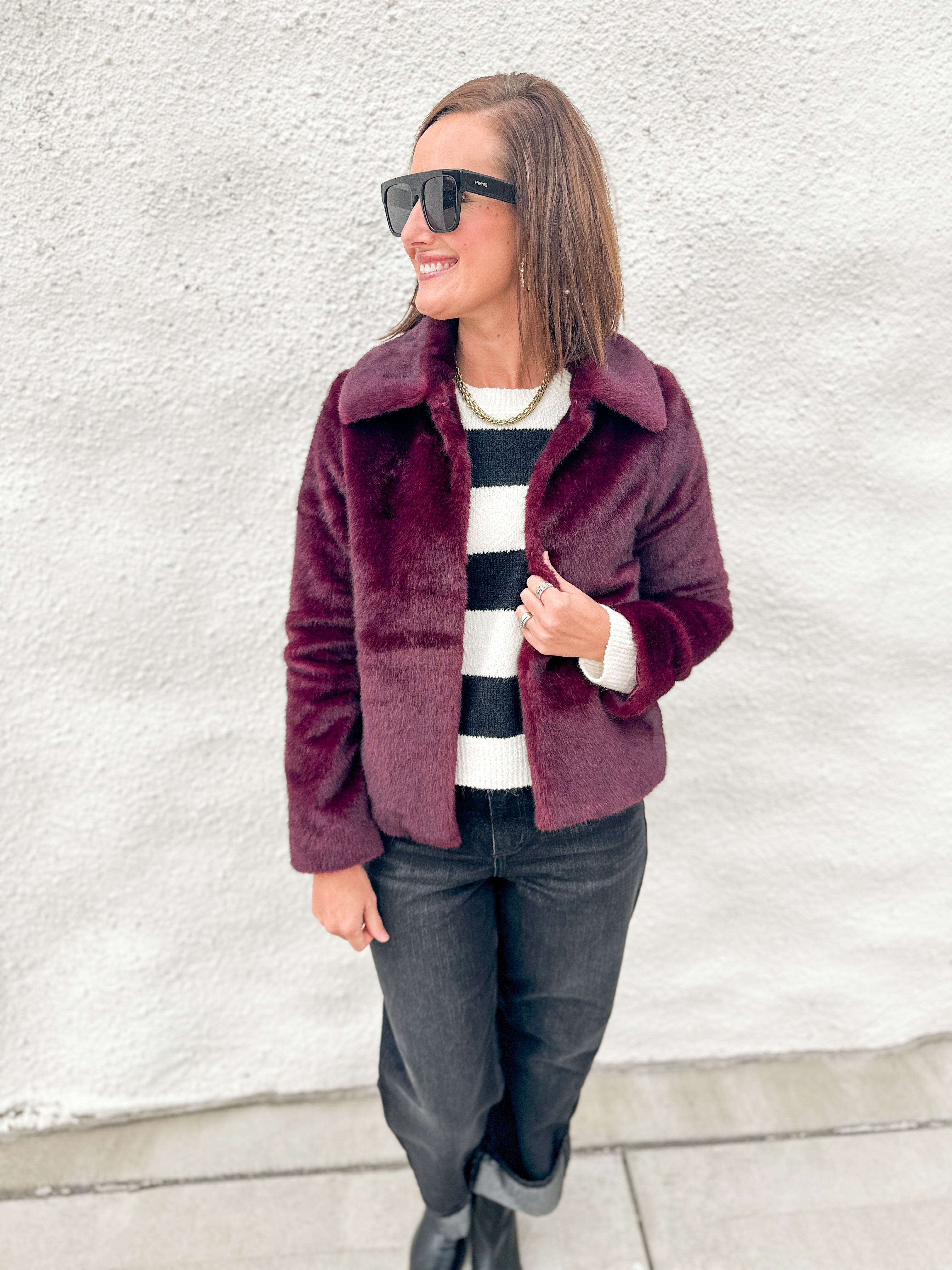 A woman wearing a burgundy faux fur jacket over a black and white striped sweater, dark jeans, and black boots, standing against a white textured wall.