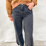 A person wearing dark charcoal gray wide-leg cargo jeans, a tan ribbed knit sweater, and brown Mary Jane sandals, standing against a white textured wall.