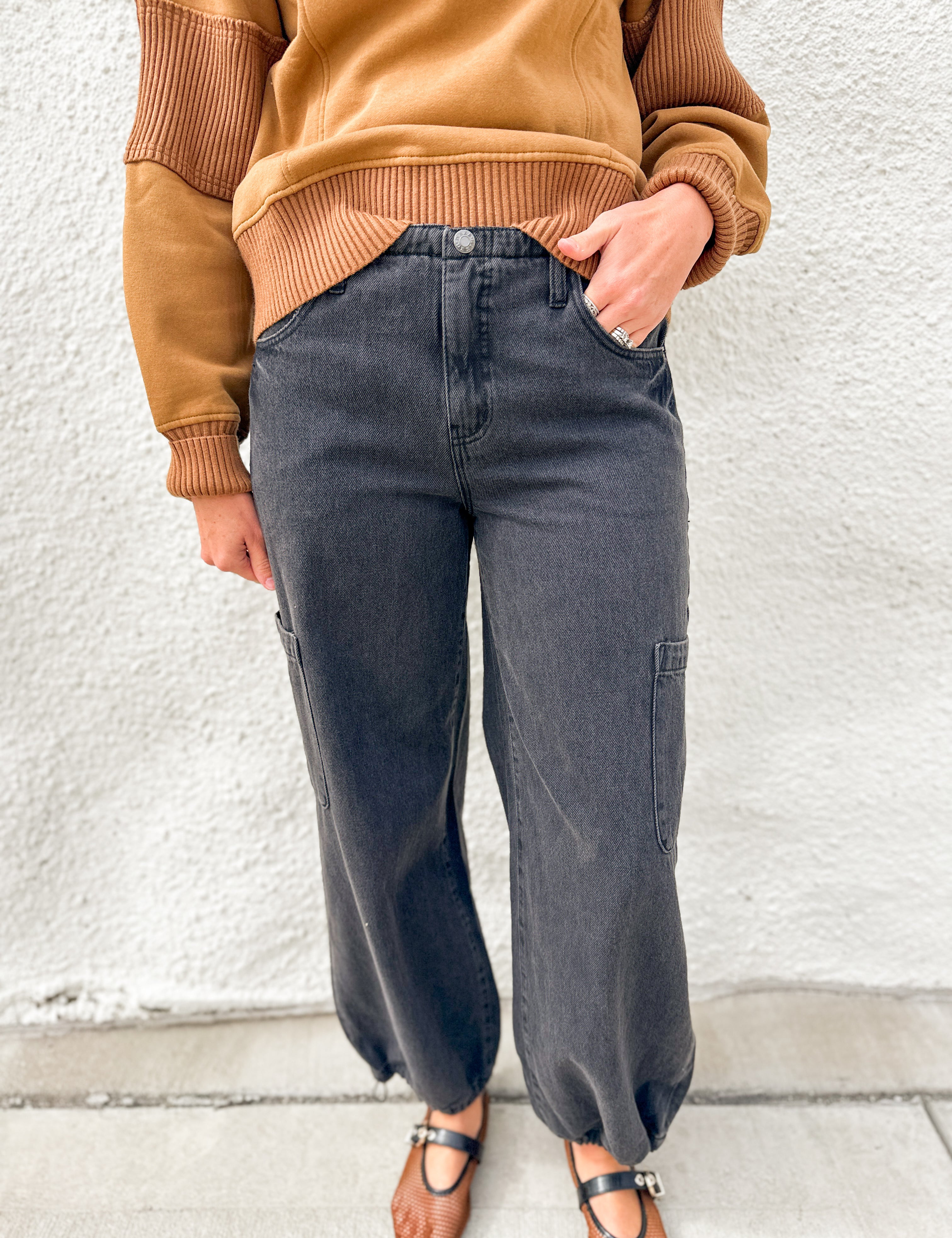 A person wearing dark charcoal gray wide-leg cargo jeans, a tan ribbed knit sweater, and brown Mary Jane sandals, standing against a white textured wall.