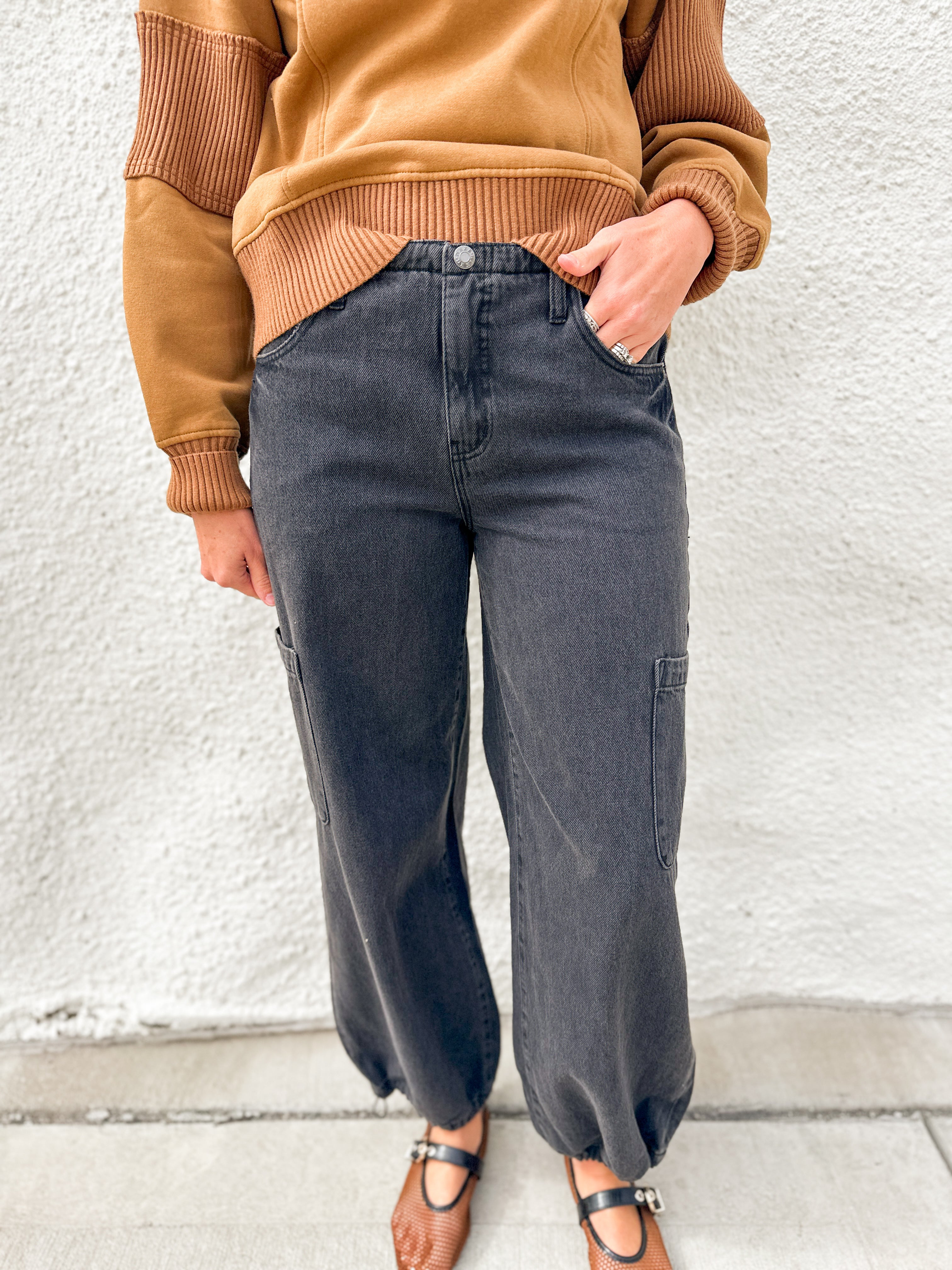 A person wearing dark charcoal gray wide-leg cargo jeans, a tan ribbed knit sweater, and brown Mary Jane sandals, standing against a white textured wall.