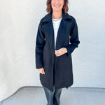 A woman stands outdoors against a light textured wall wearing a long black coat over a white top and gray pants.